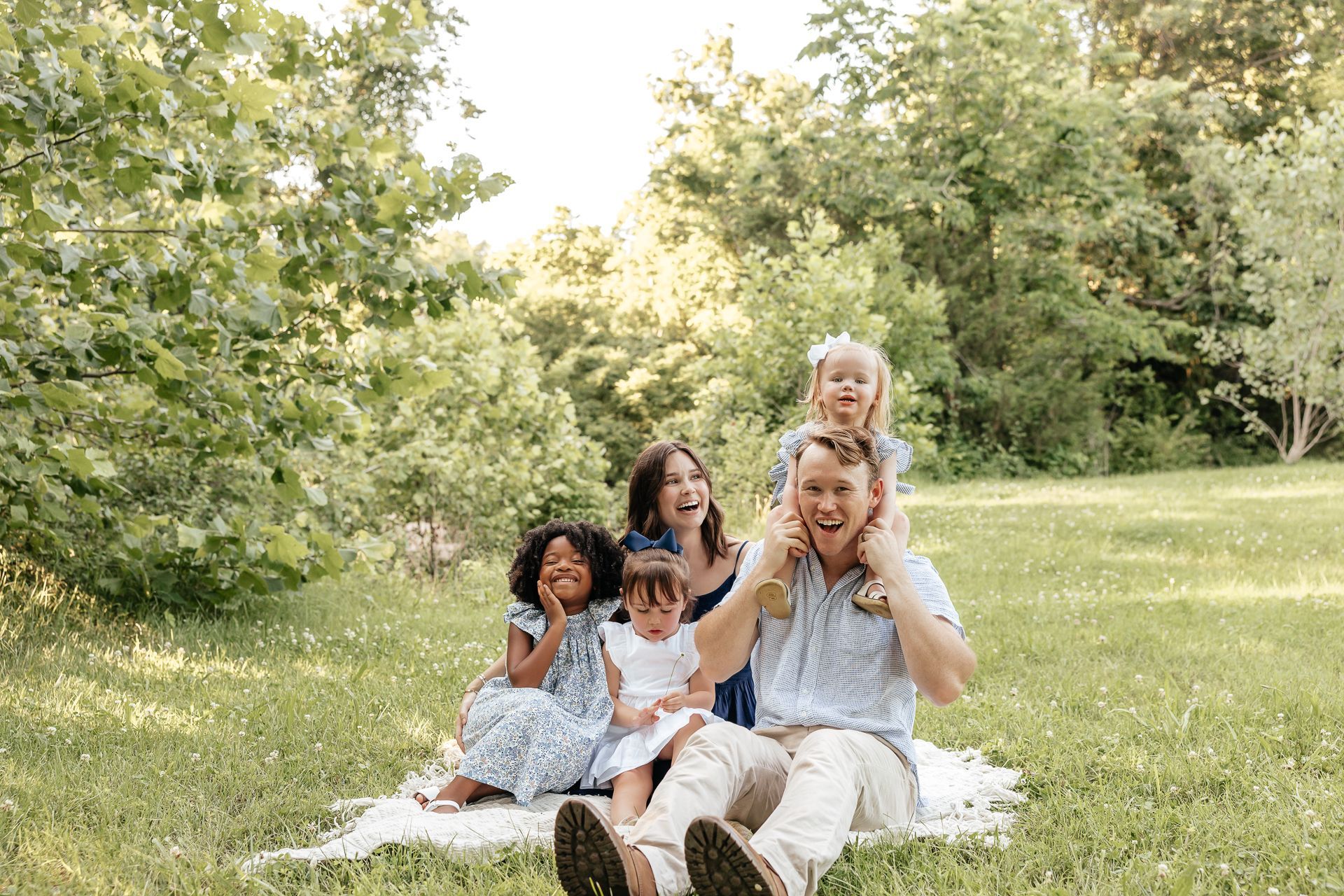 Family of five smiles on a grassy picnic blanket in a park, with trees in the background.
