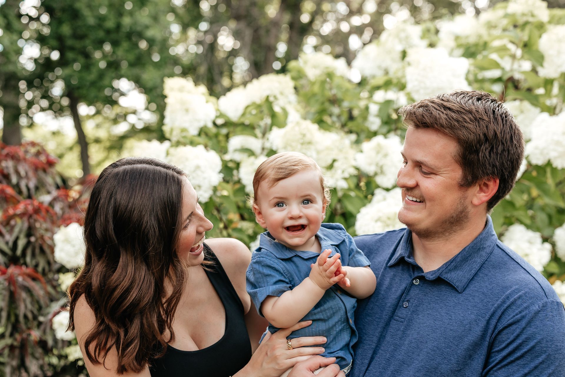 A smiling family of three poses outdoors: parents with a baby boy.