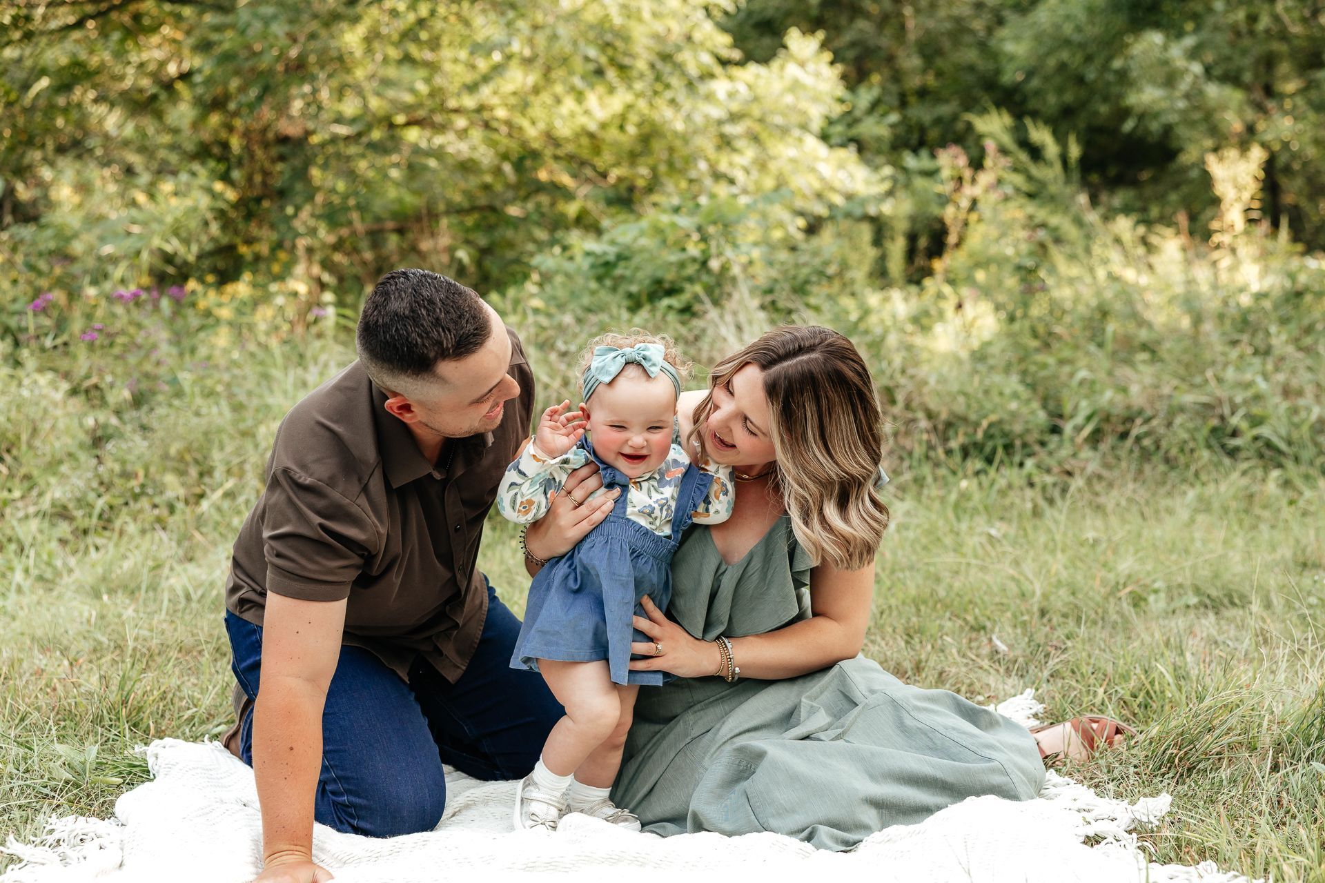 Family of three in a grassy field; parents smiling at a toddler, all on a blanket.