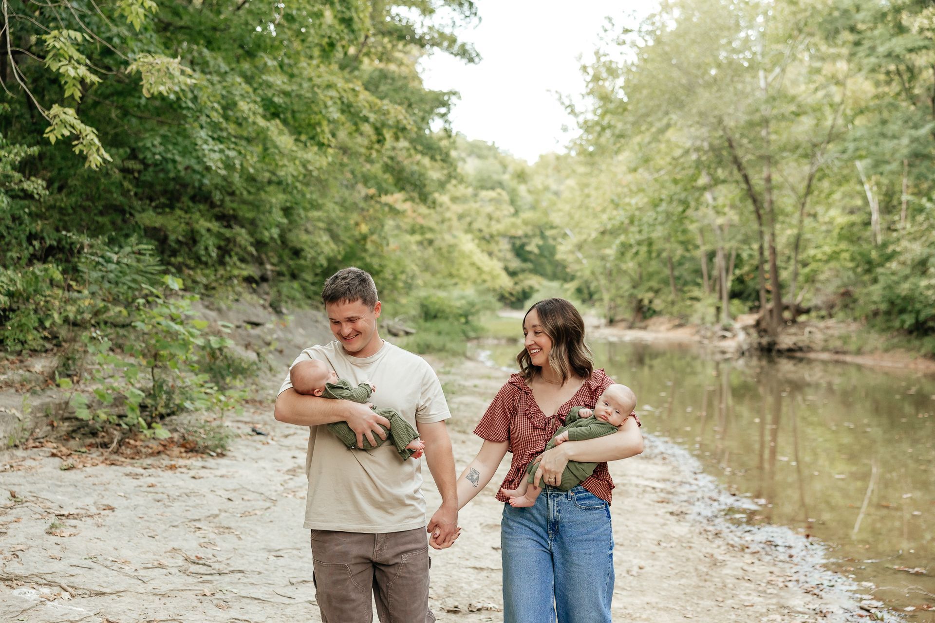 Parents holding newborn twins walk along a riverbank path; the family smiles.