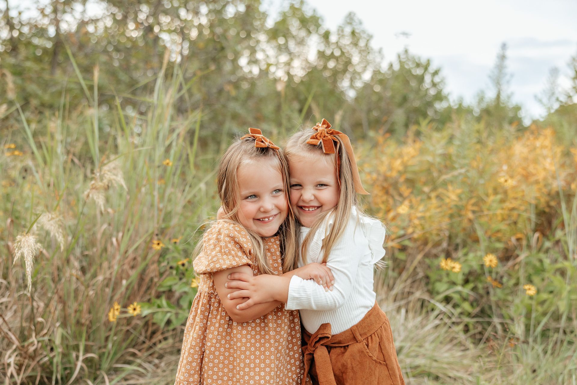 Two young girls smiling and hugging outdoors, wearing matching hair bows and fall-colored outfits.