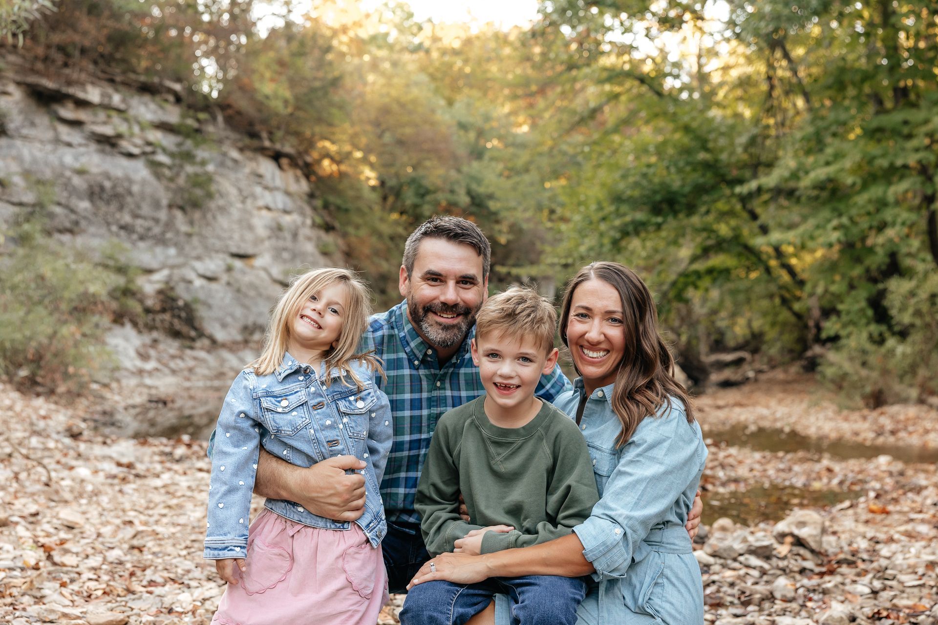 Family of four smiling, posing outdoors by a riverbed with trees and rocky cliff.