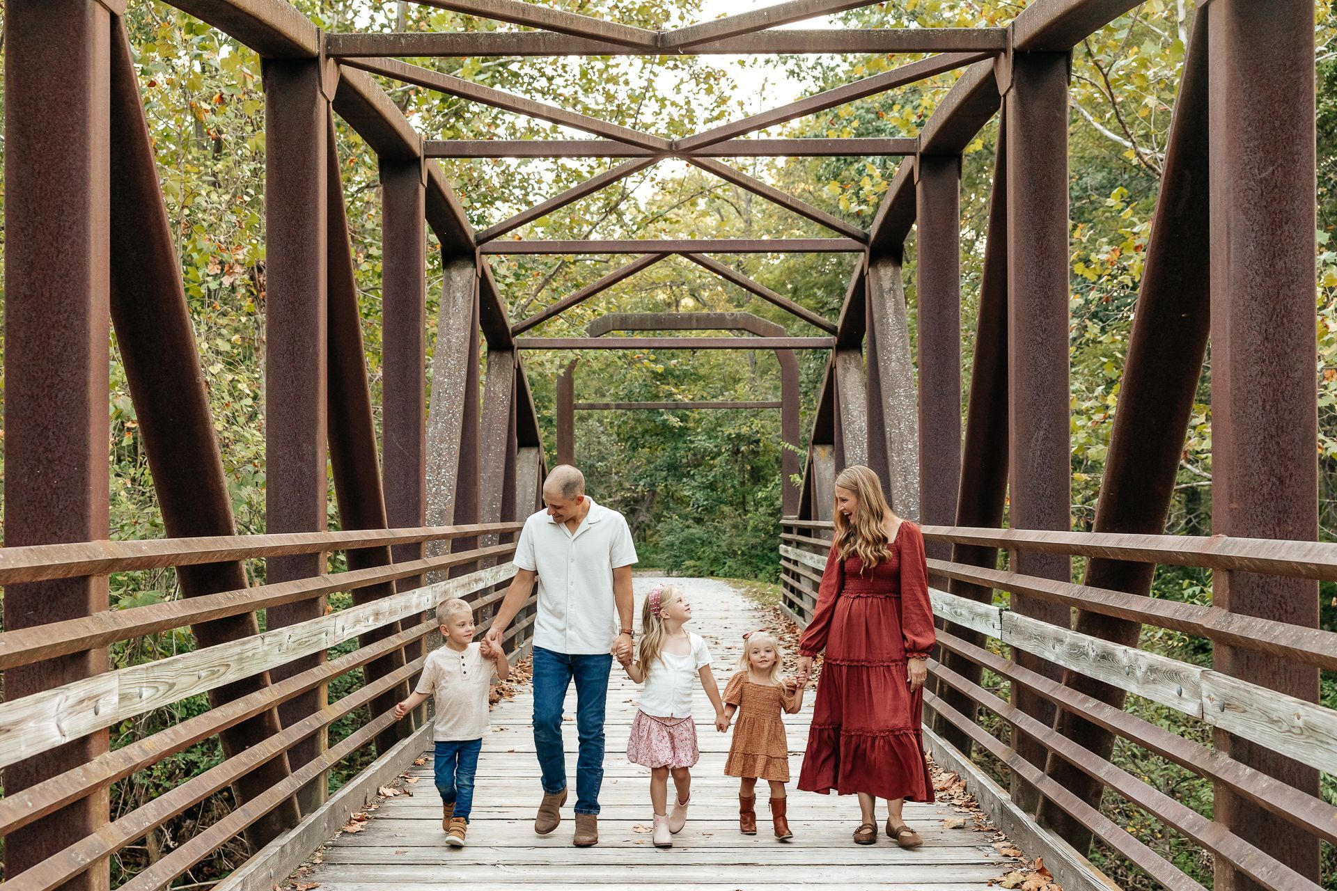 Family walking hand-in-hand on a rusty bridge, framed by metal beams and surrounded by trees.