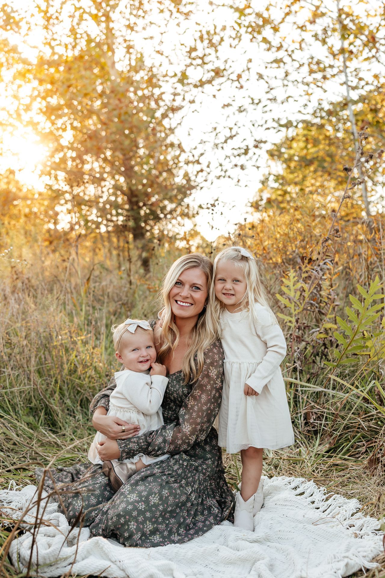 Woman and two young children smiling in a sunlit field, sitting on a blanket.