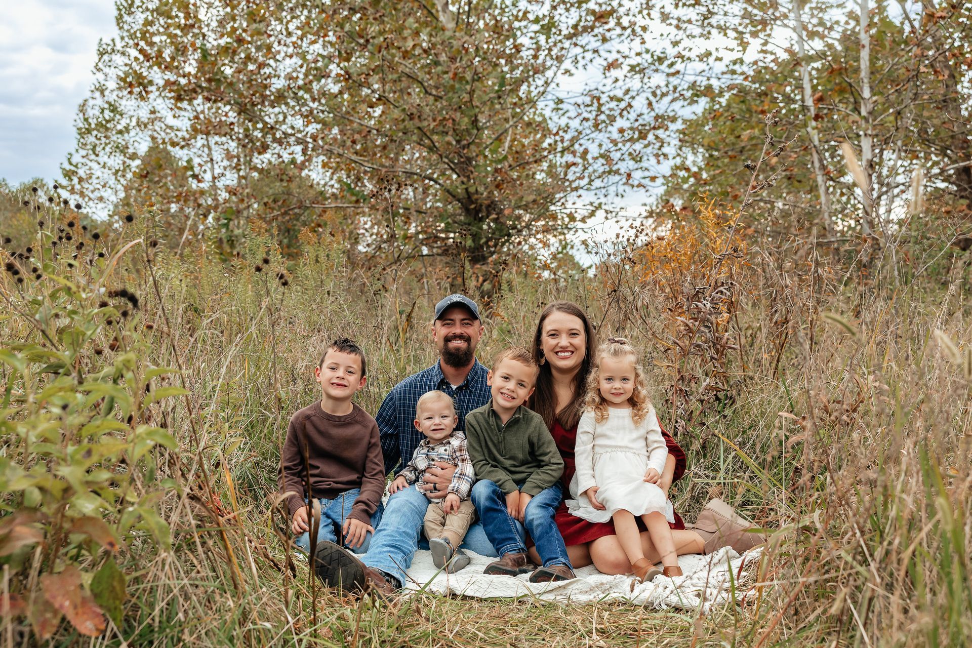 Family of six sits on a blanket in tall grass; fall foliage in the background.