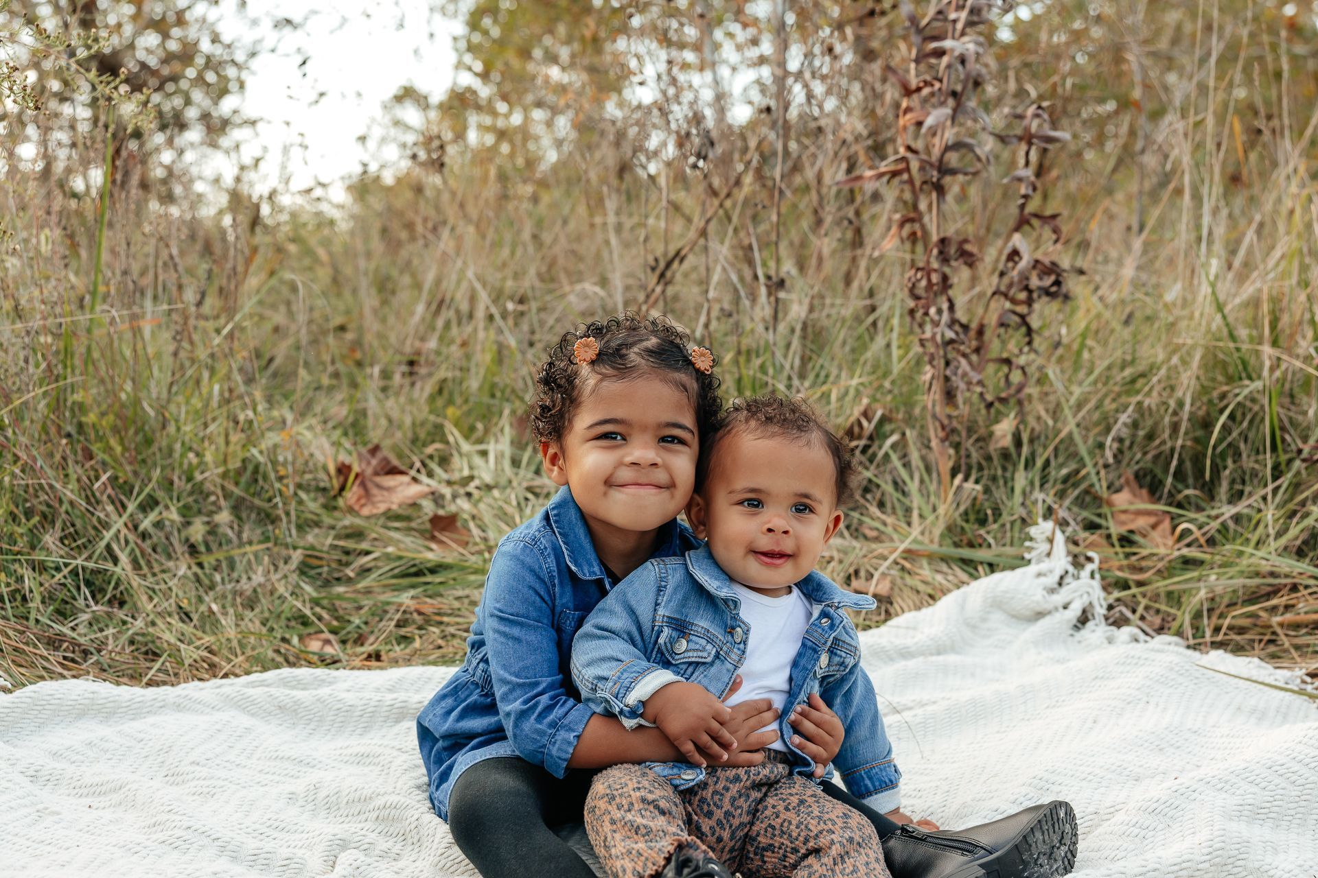 Two children, one in a denim jacket, hug on a blanket in a field, smiling.