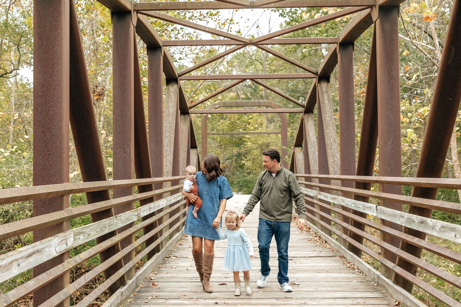 Family of four walking hand-in-hand on a wooden bridge, autumn foliage in the background.