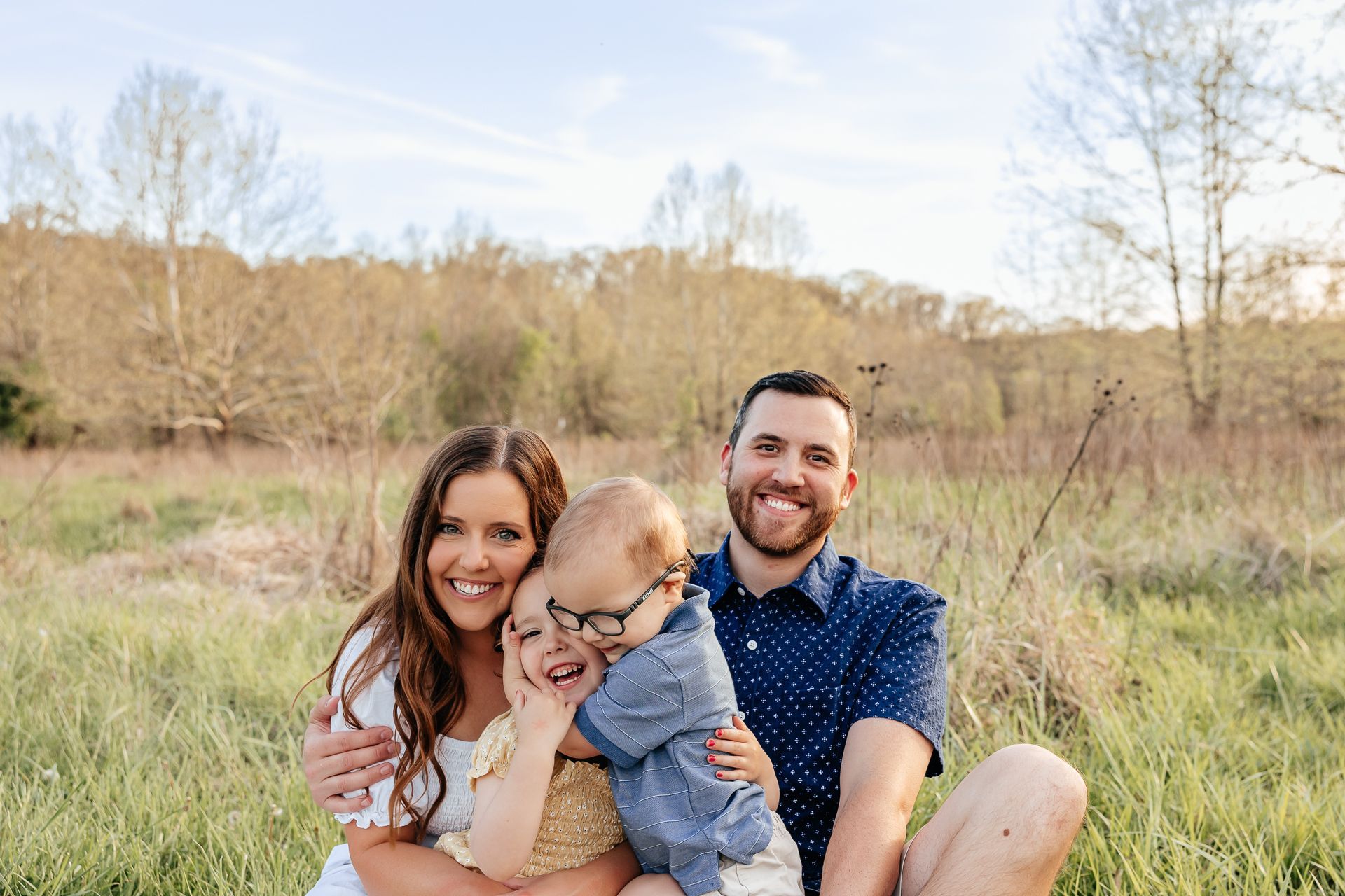 Family of four smiling in a grassy field. Mother and father embrace two small children.