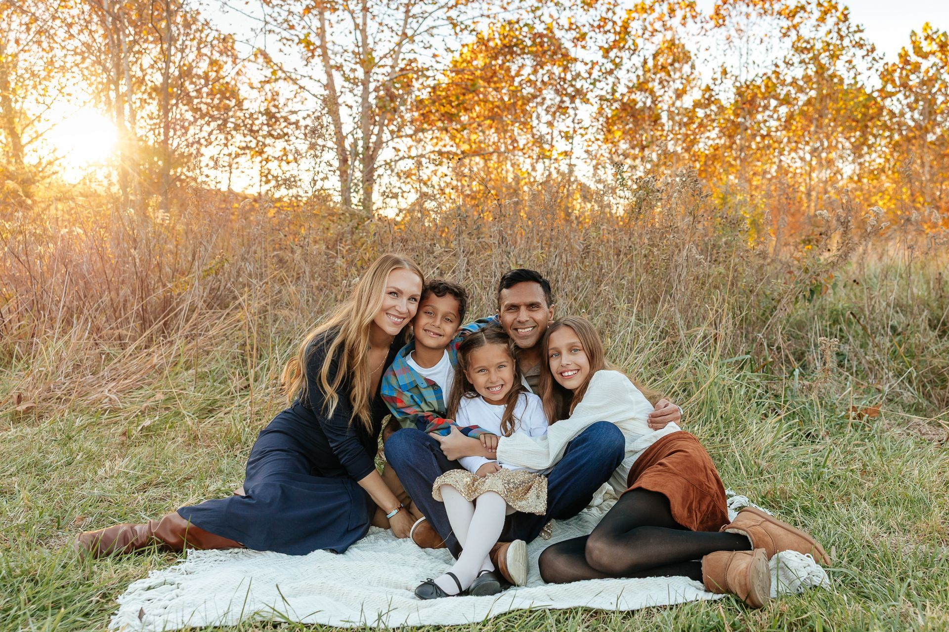 Family sitting in a field, smiling at the camera. Sunlight, fall foliage, blanket.