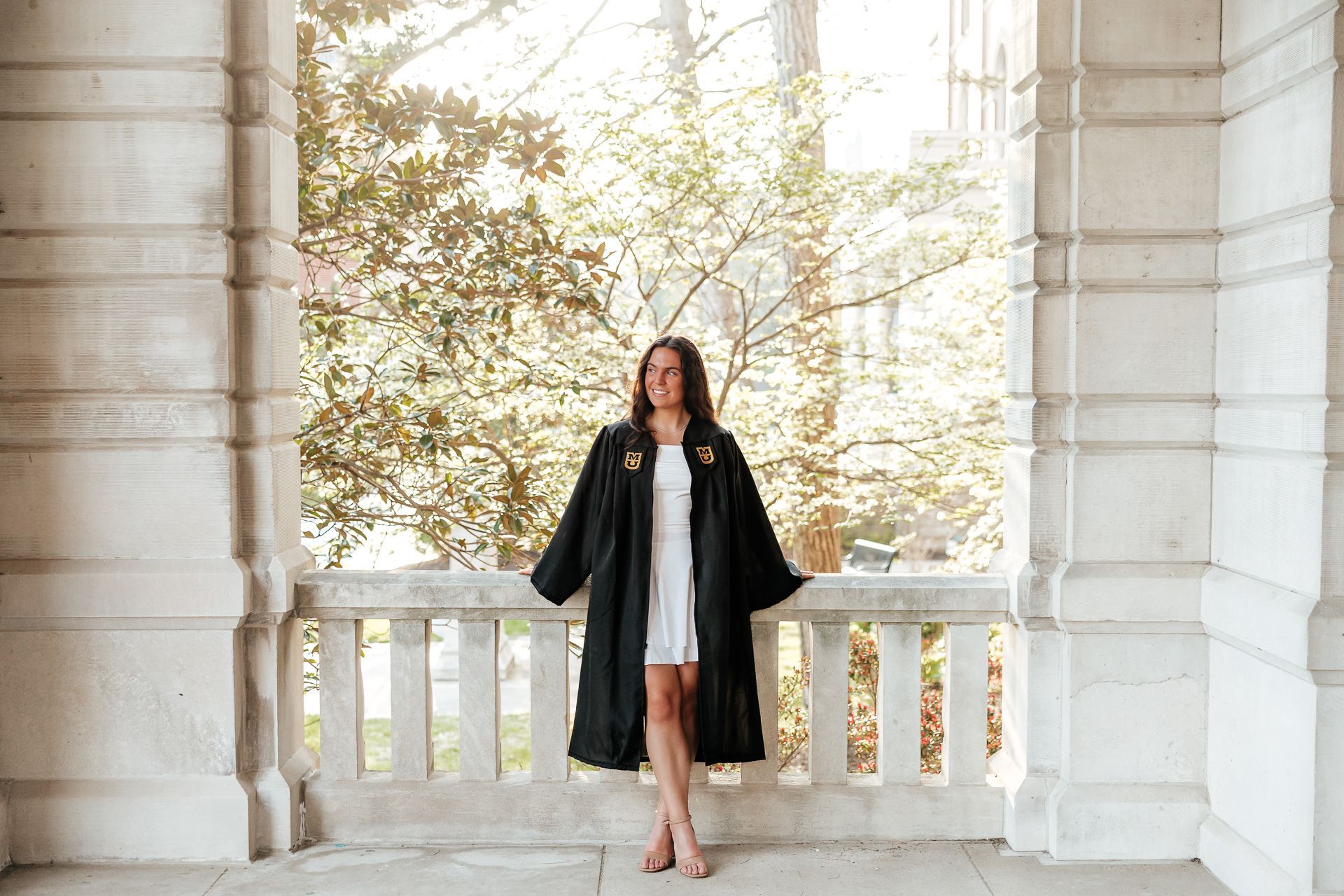 Woman in graduation gown stands on a balcony, smiling. Sunlight filters through trees.