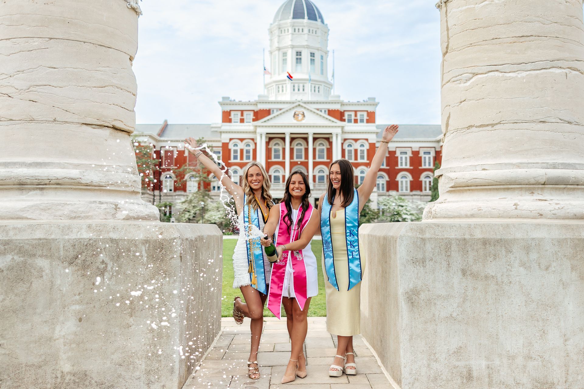 Three graduates celebrate, throwing confetti, in front of a large building with a dome.