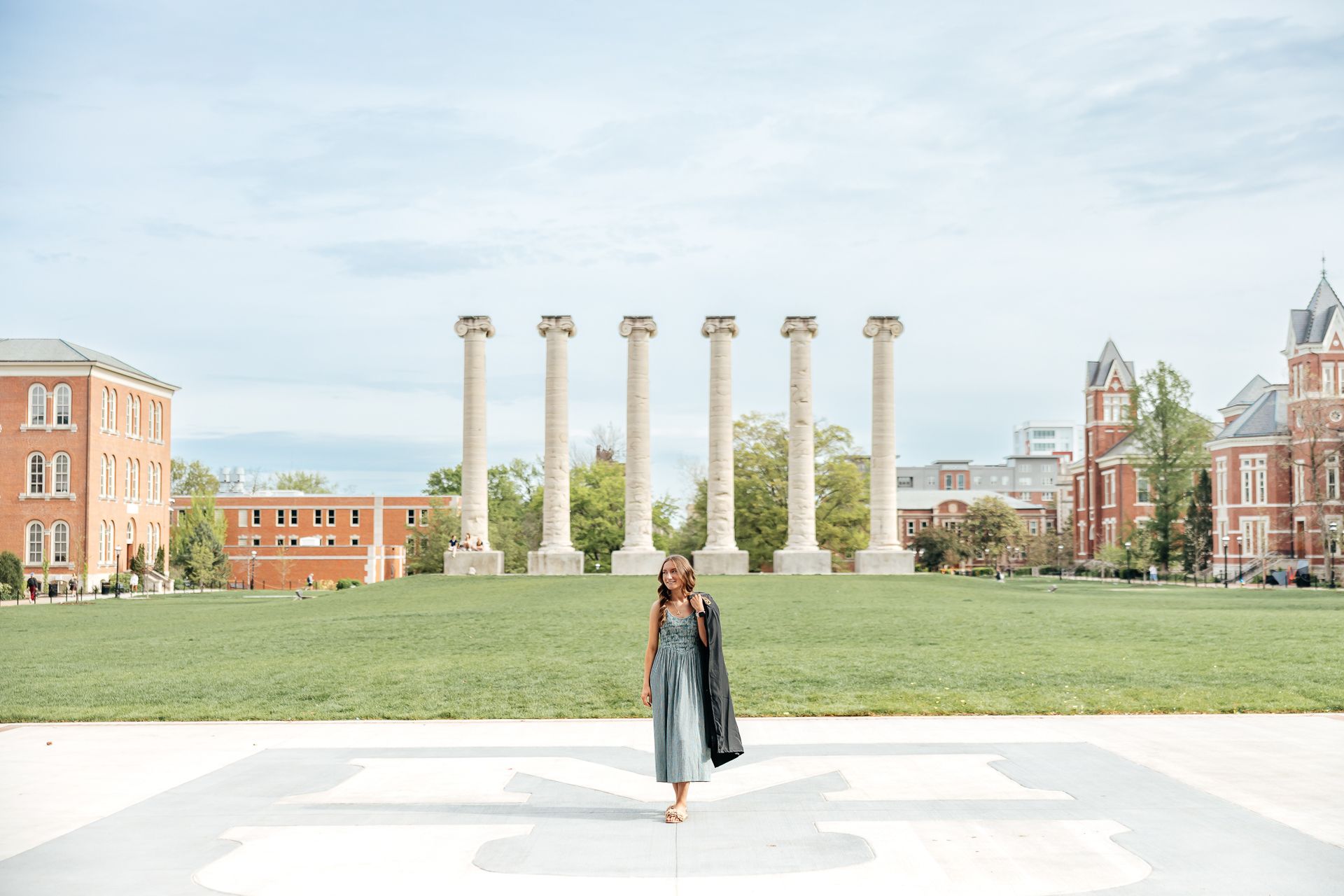 Woman standing on a large concrete M in front of six stone columns on a green lawn, buildings in the background.