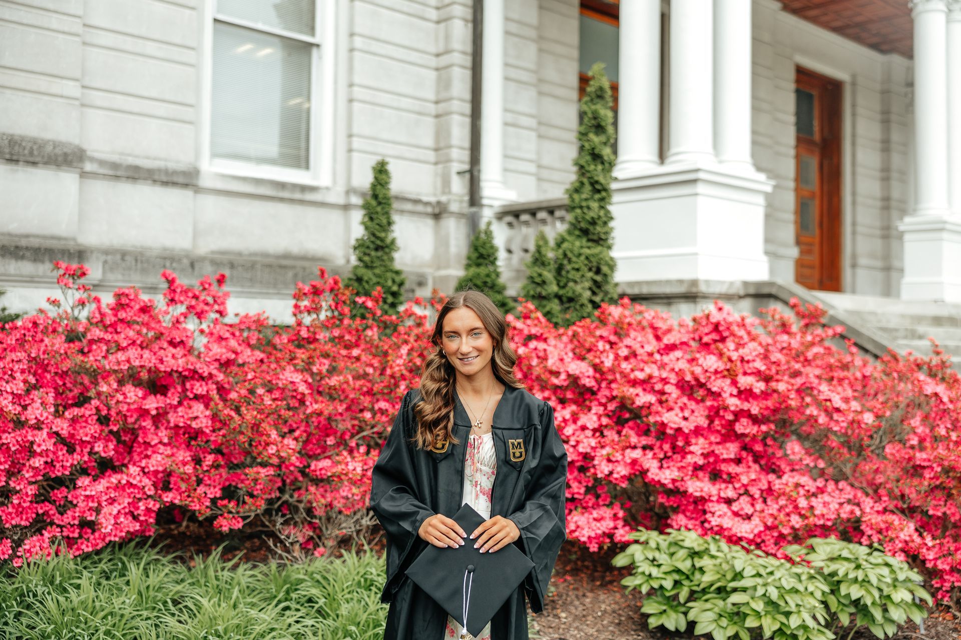 Graduation portrait: woman in cap and gown, smiling, holding cap in front of building with columns and red bushes.