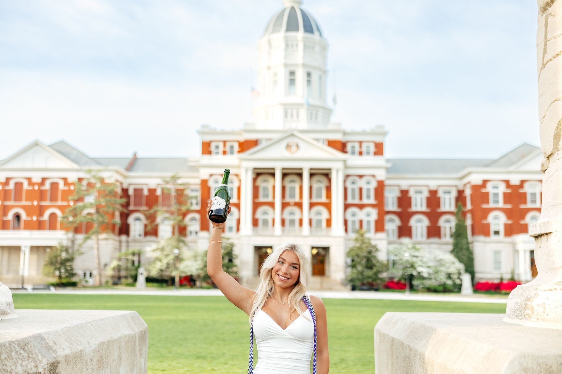 Woman in graduation gown holding champagne bottle in front of a university building.