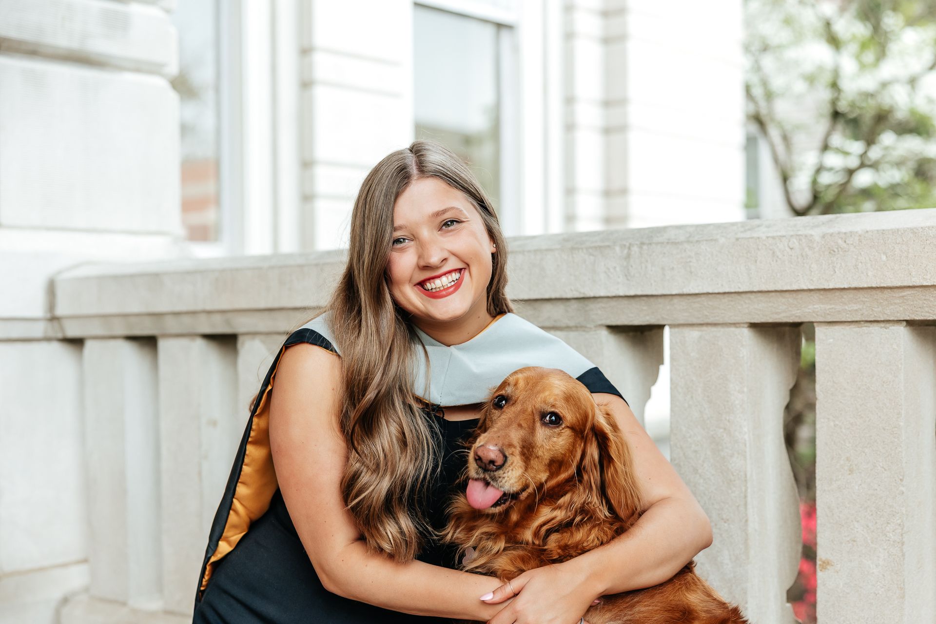 Woman in graduation attire hugging a golden retriever, smiling, outdoors.
