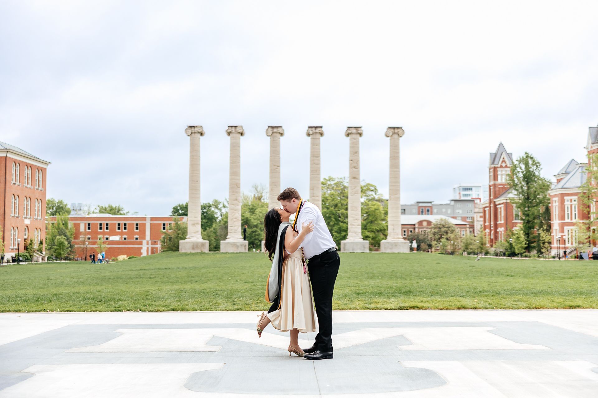 Couple kissing in front of tall columns on a green lawn, buildings in background.