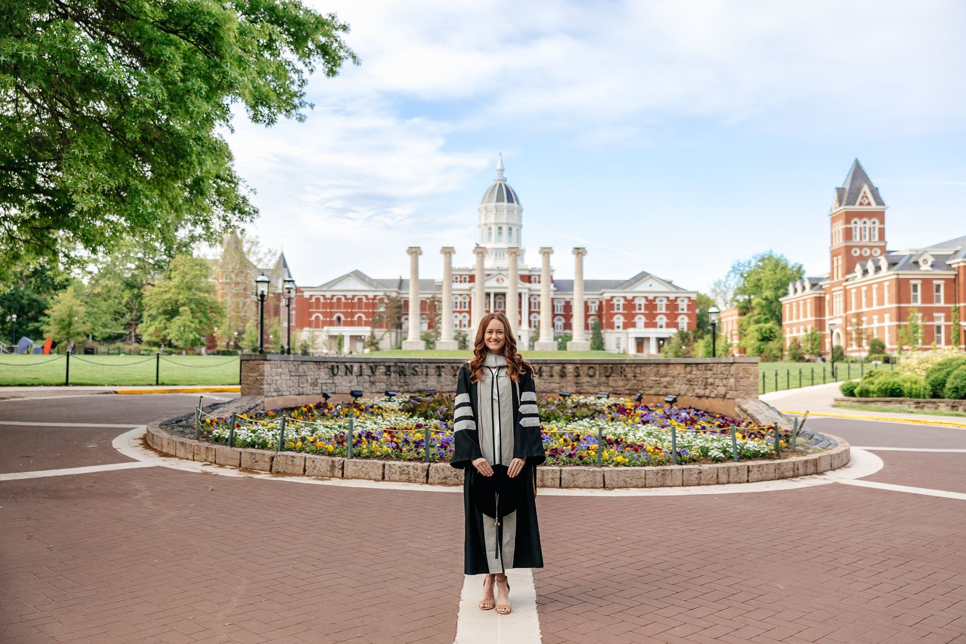 Woman in graduation gown stands in front of a campus building with flowers.