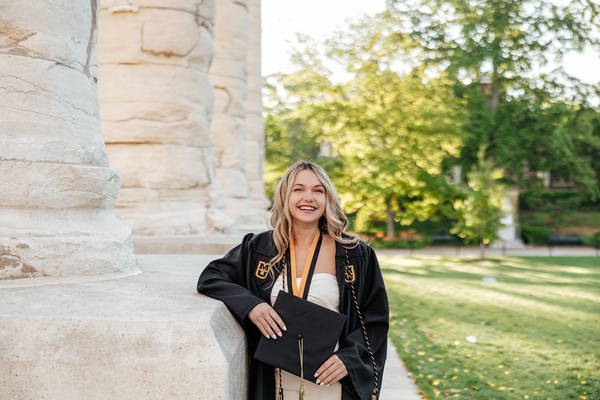 Woman in graduation gown smiles, holding a cap, leaning against a stone column. Trees in background.