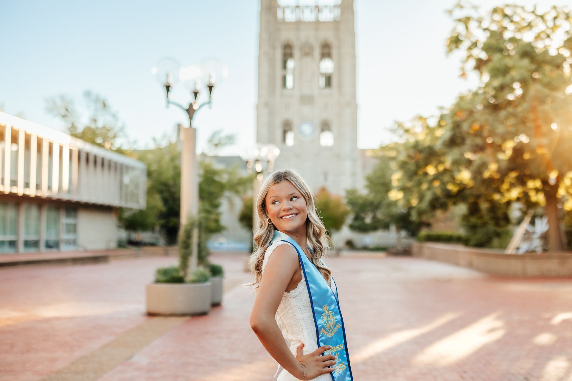 Woman in graduation gown smiles on campus, bell tower in background.