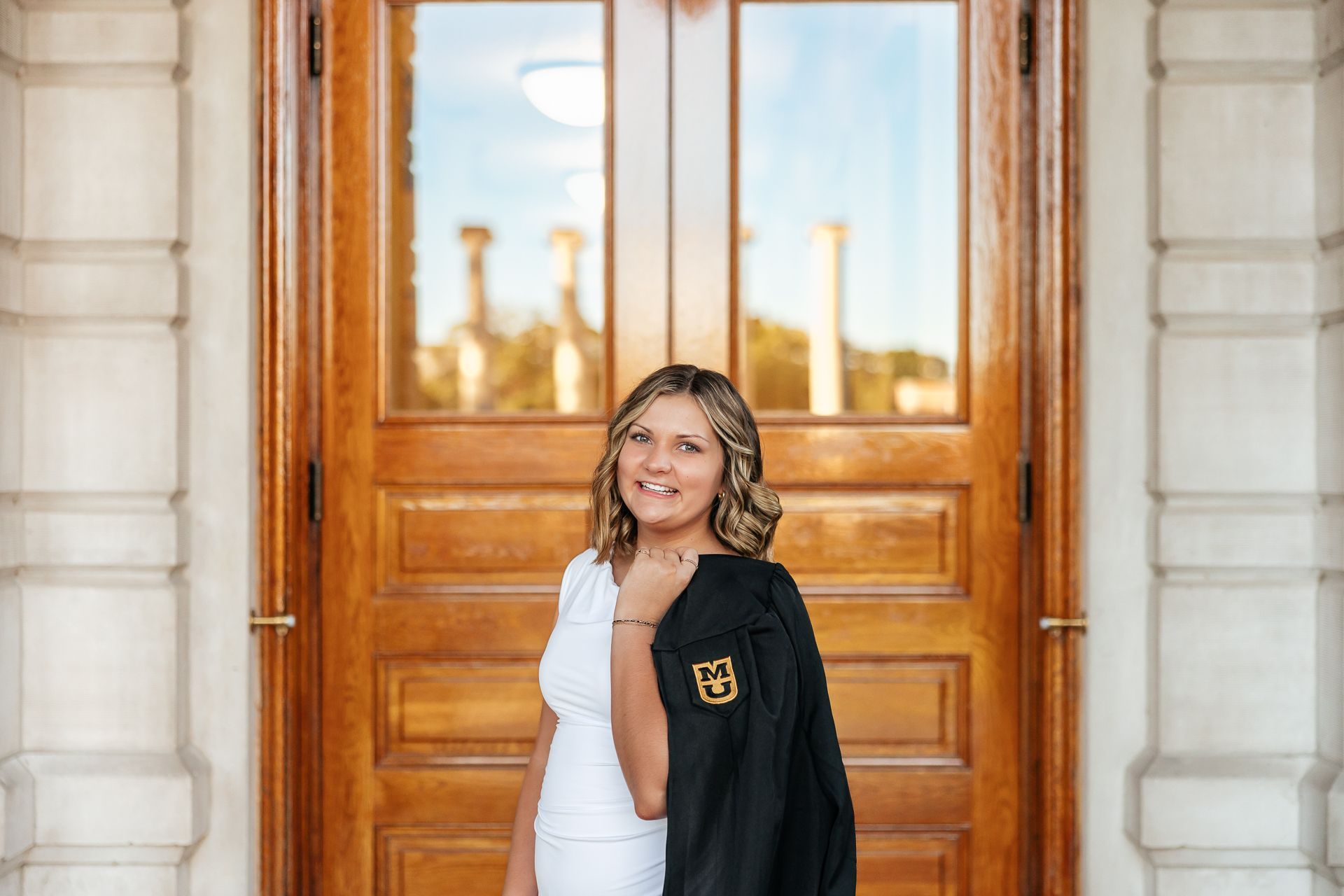 Woman in white dress smiles, holding a black jacket with a gold emblem, standing in front of large wooden doors.