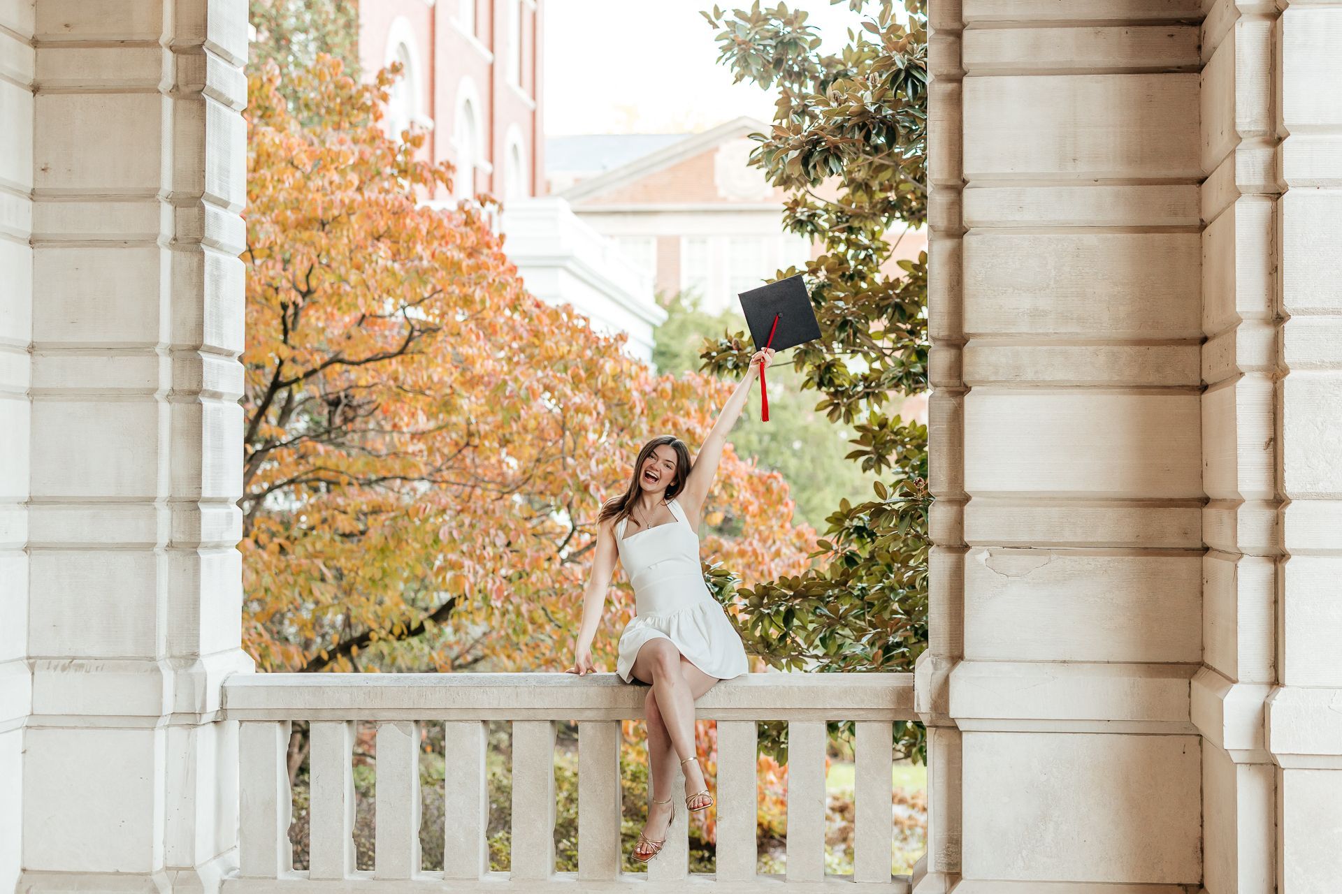 Woman celebrating graduation, raising cap, sitting on a balcony. Fall foliage, white dress, joyful expression.