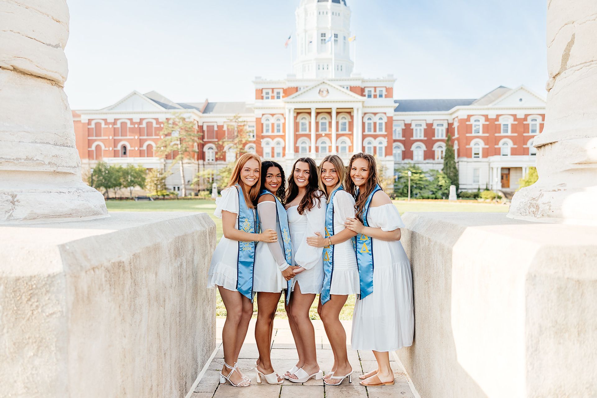 Six women in white dresses with blue stoles pose in front of a brick building.