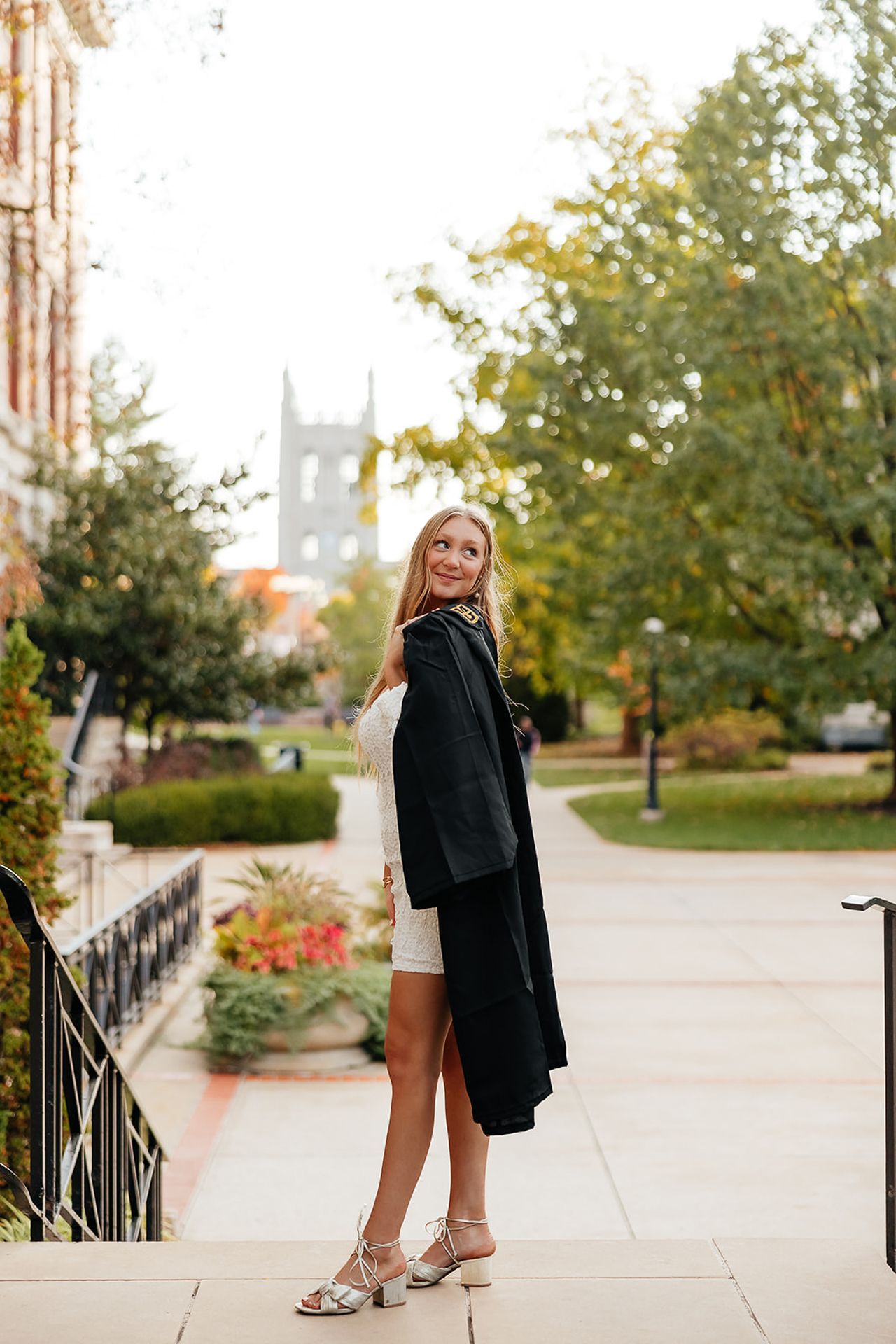 Woman in white dress and black coat poses on steps, with a university building in the background.