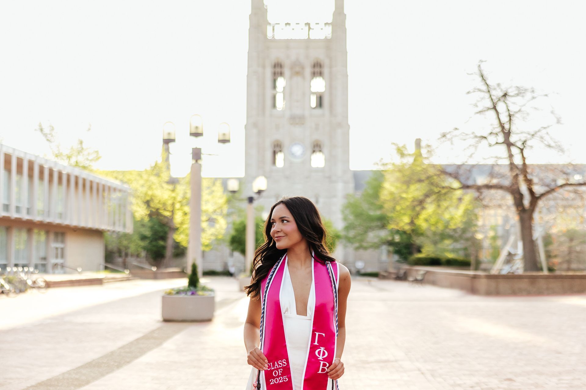 Woman wearing a pink sash smiles in front of a university building.