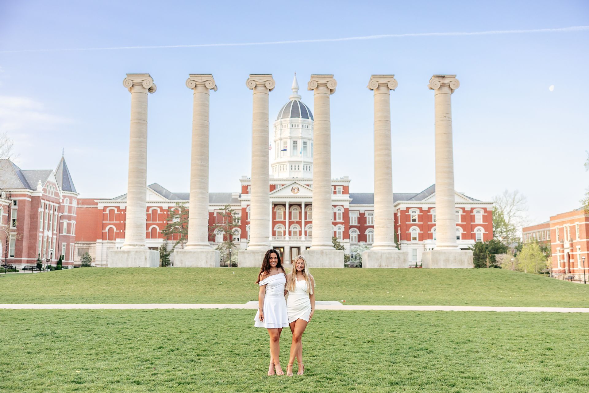 Two women in white dresses pose in front of the columns and main building on the University of Missouri campus.