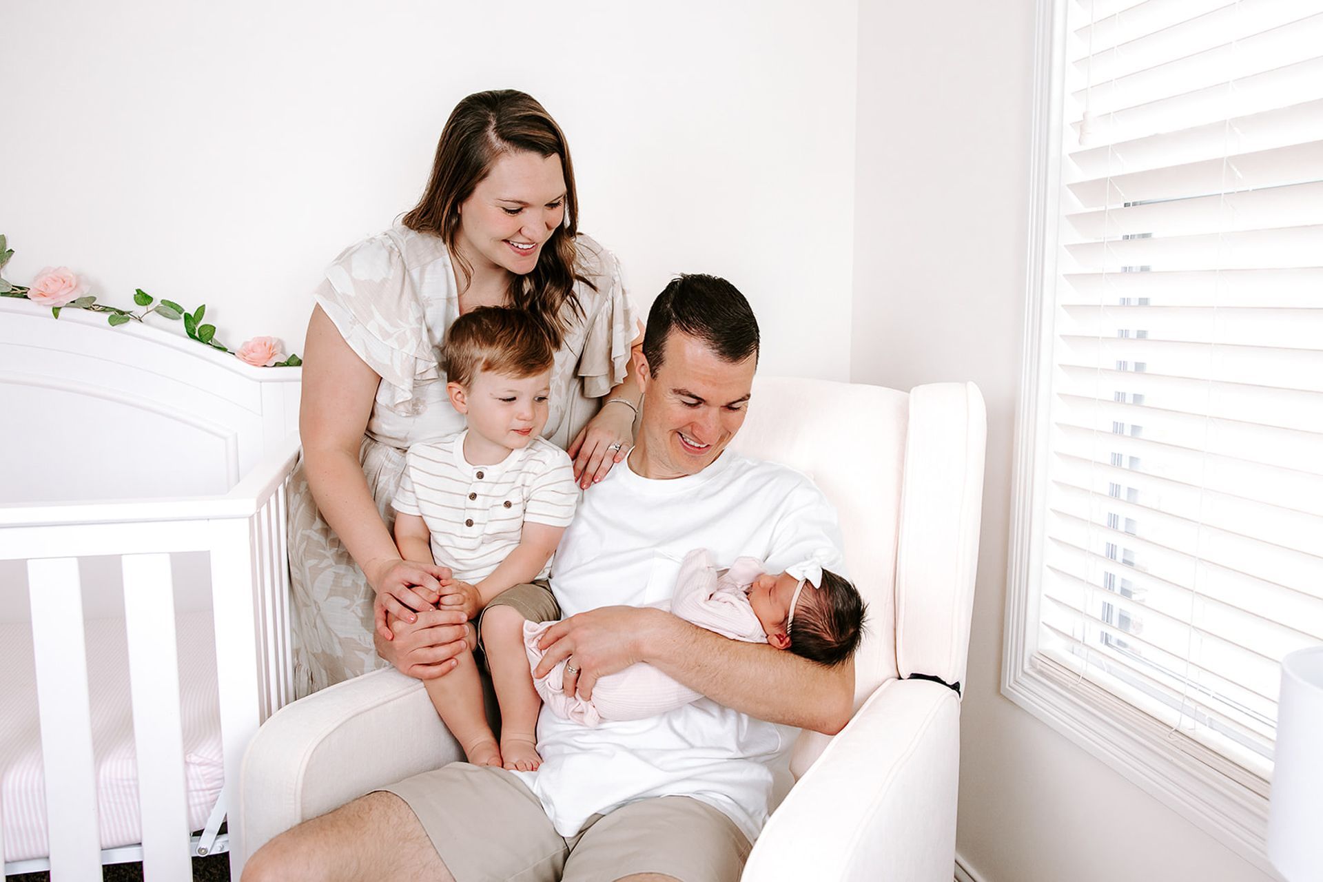 Family of four in nursery: Father holding newborn, mother and older child watching, white crib and glider.