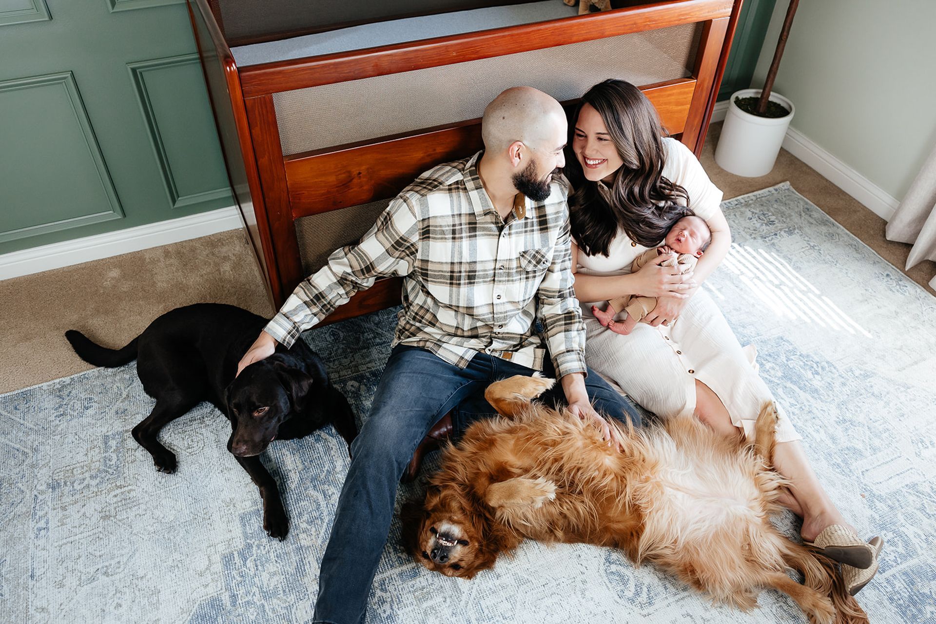 Couple with newborn, two dogs, on rug near crib. Smiling parents, holding baby.