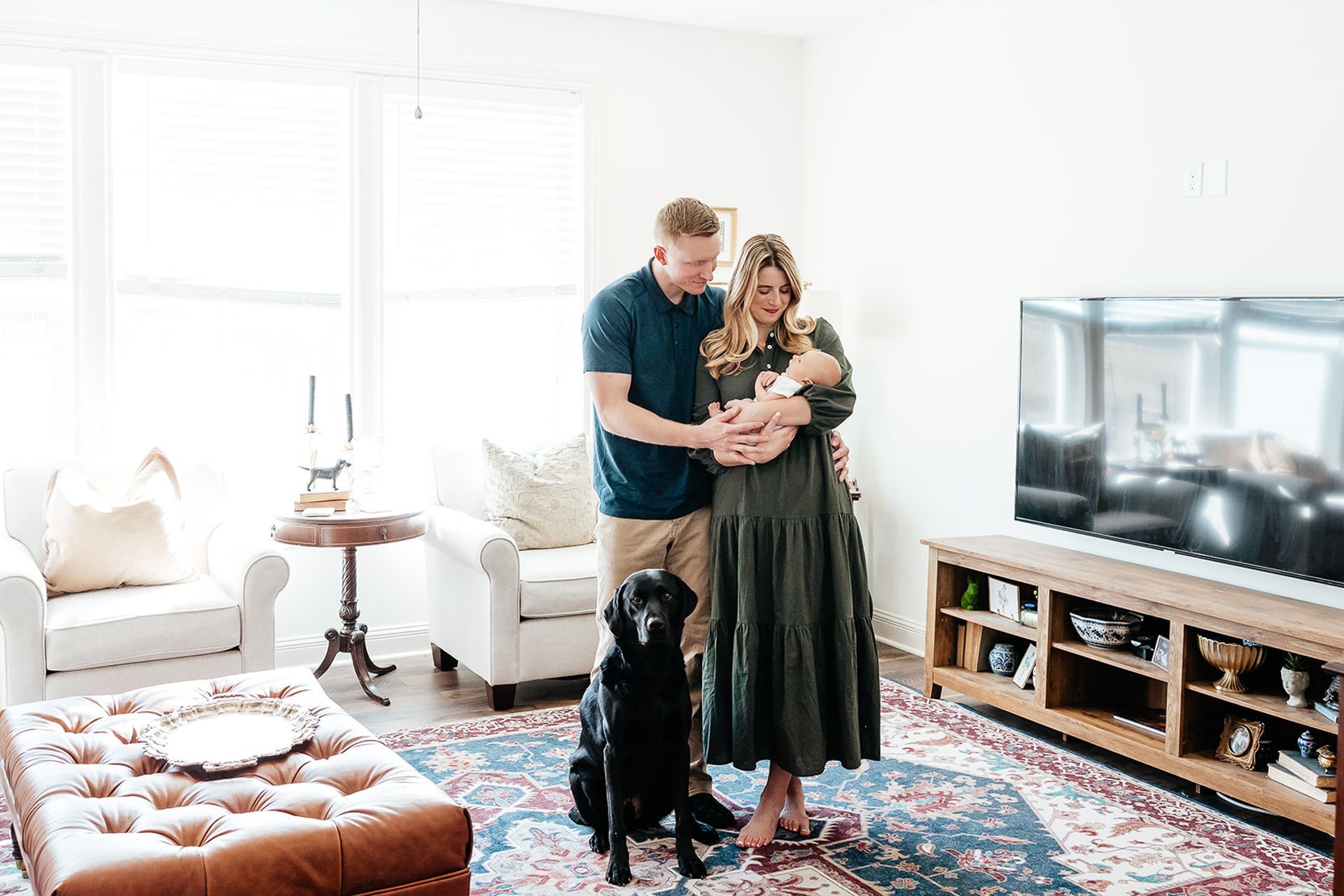 Parents with newborn and dog in a bright living room. Woman holds baby; man touches them.