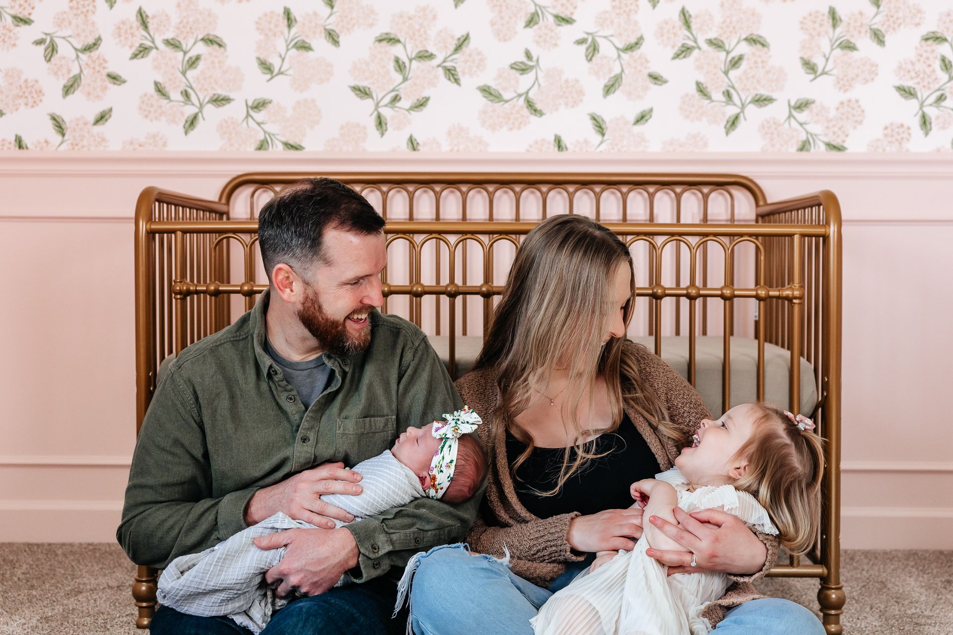 Family of four with a newborn and toddler, smiling. Pink and gold nursery backdrop.