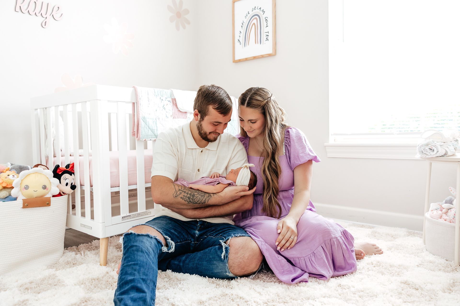 Parents seated on a rug hold their newborn in a nursery with a crib and rainbow art.