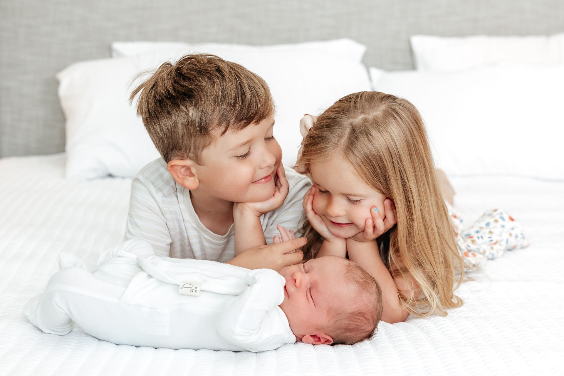 Two children smile at a newborn baby lying on a white bed.