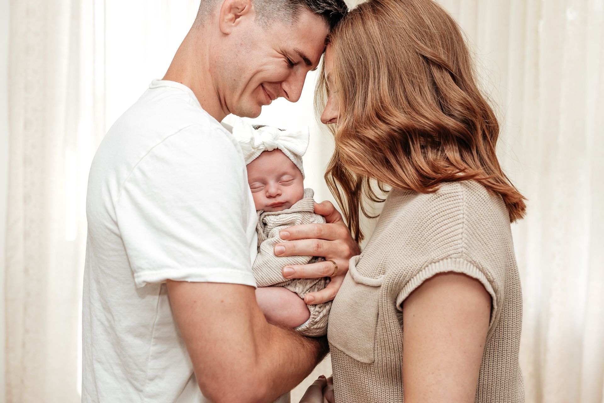 Parents gently hold a newborn baby, forehead to forehead, near sheer curtains.