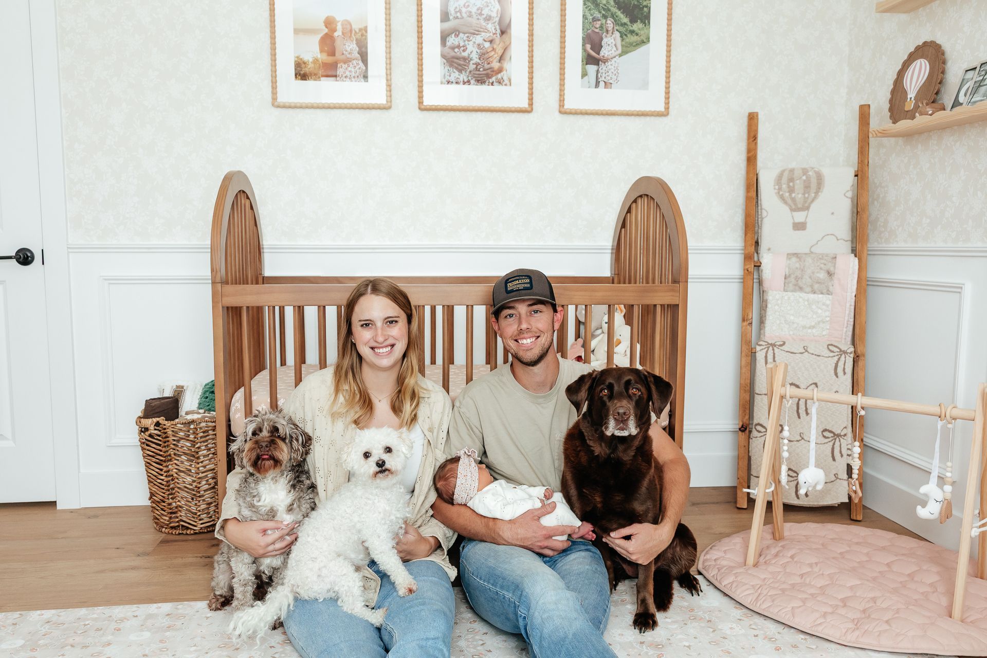 Family with newborn and dogs in nursery; smiling, sitting. Wooden crib, artwork, neutral colors.