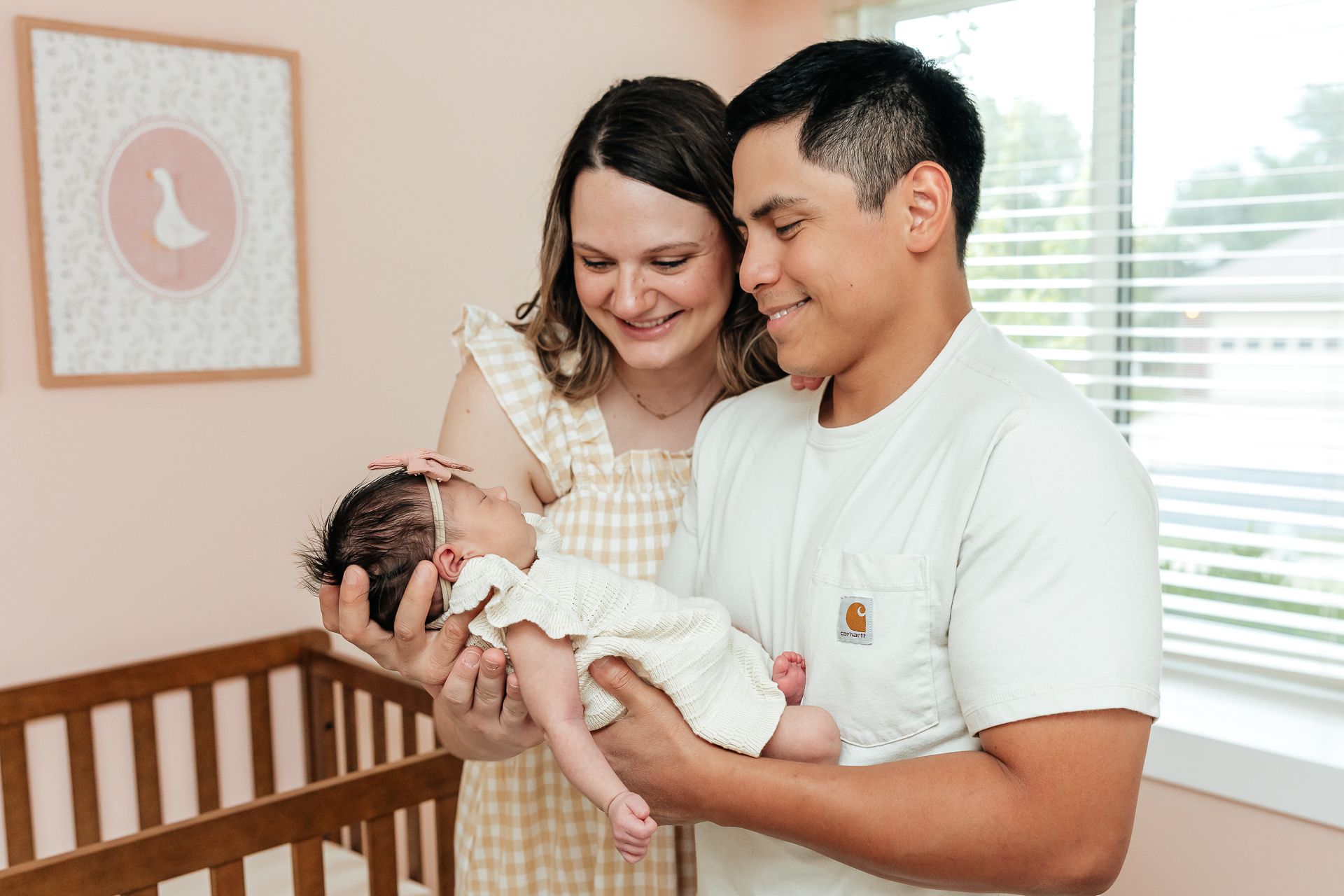 Parents holding newborn in nursery, smiling. Pink wall, framed artwork, crib visible.