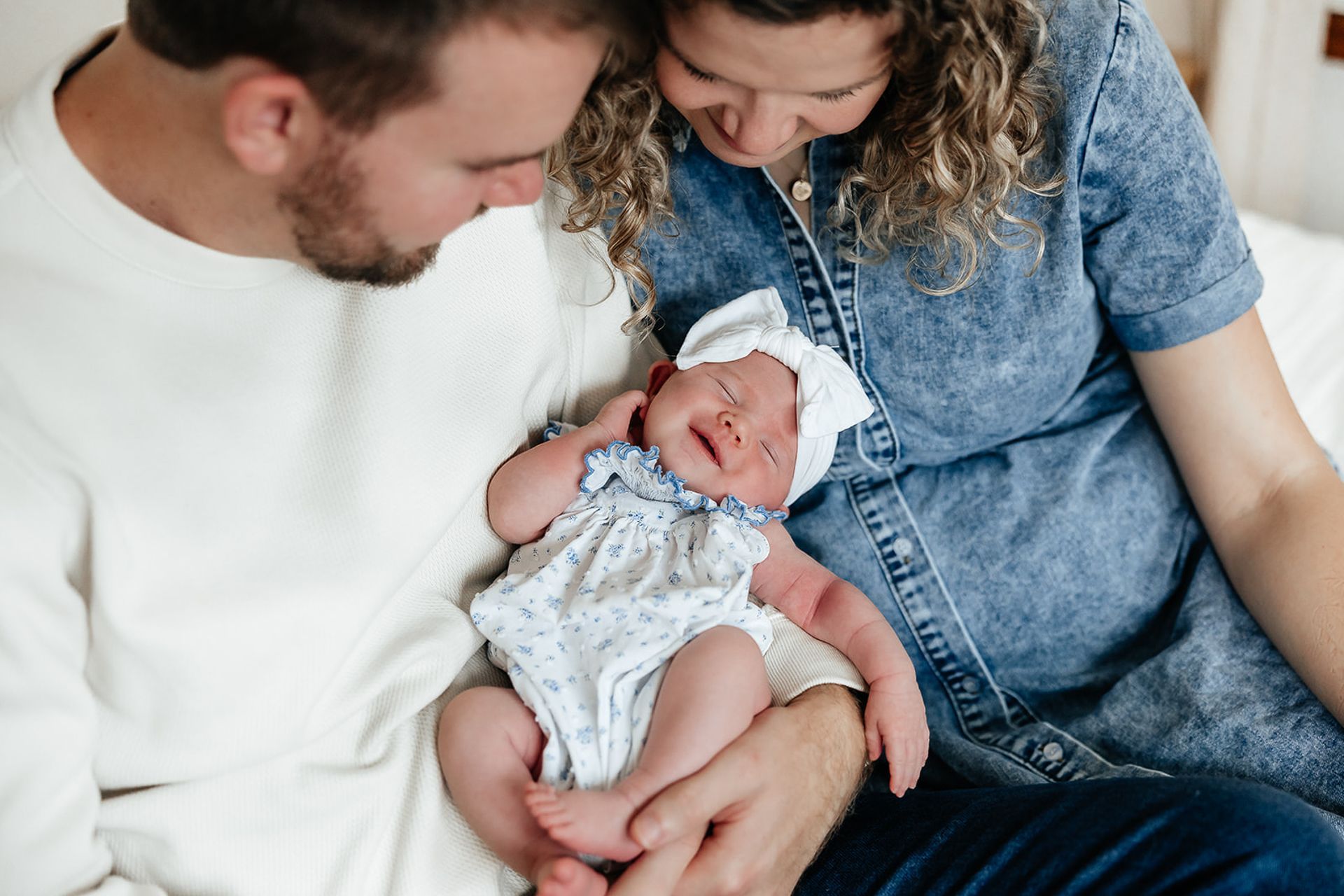 Parents holding newborn baby, all smiling. Baby wears a white bow.