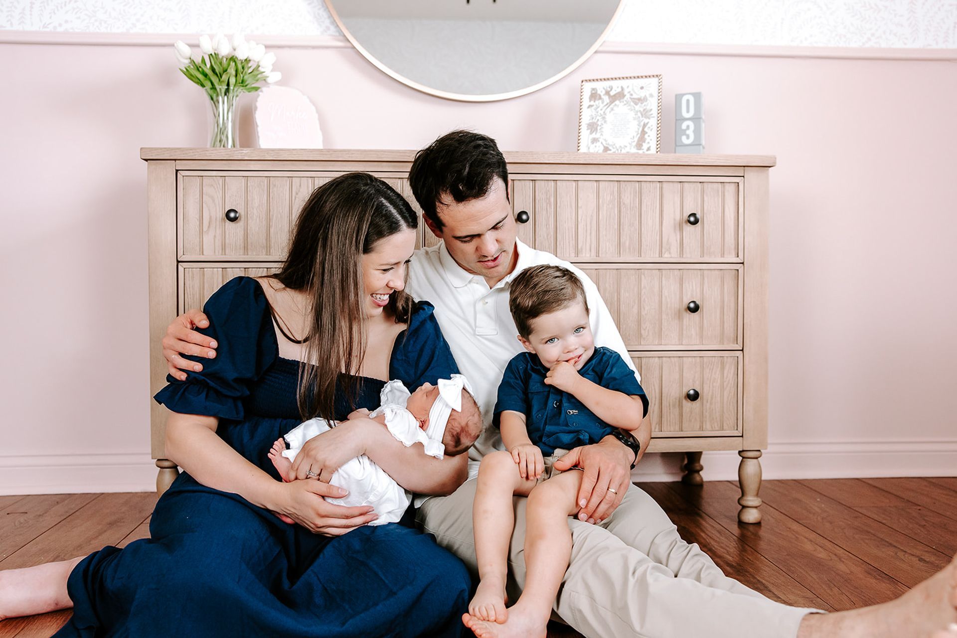 Family sits on the floor with newborn, toddler, and dresser in a pink room.