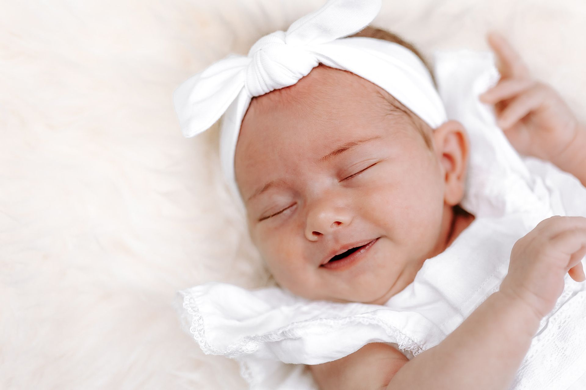 Newborn wearing a white bow headband, sleeping on a fluffy white surface.