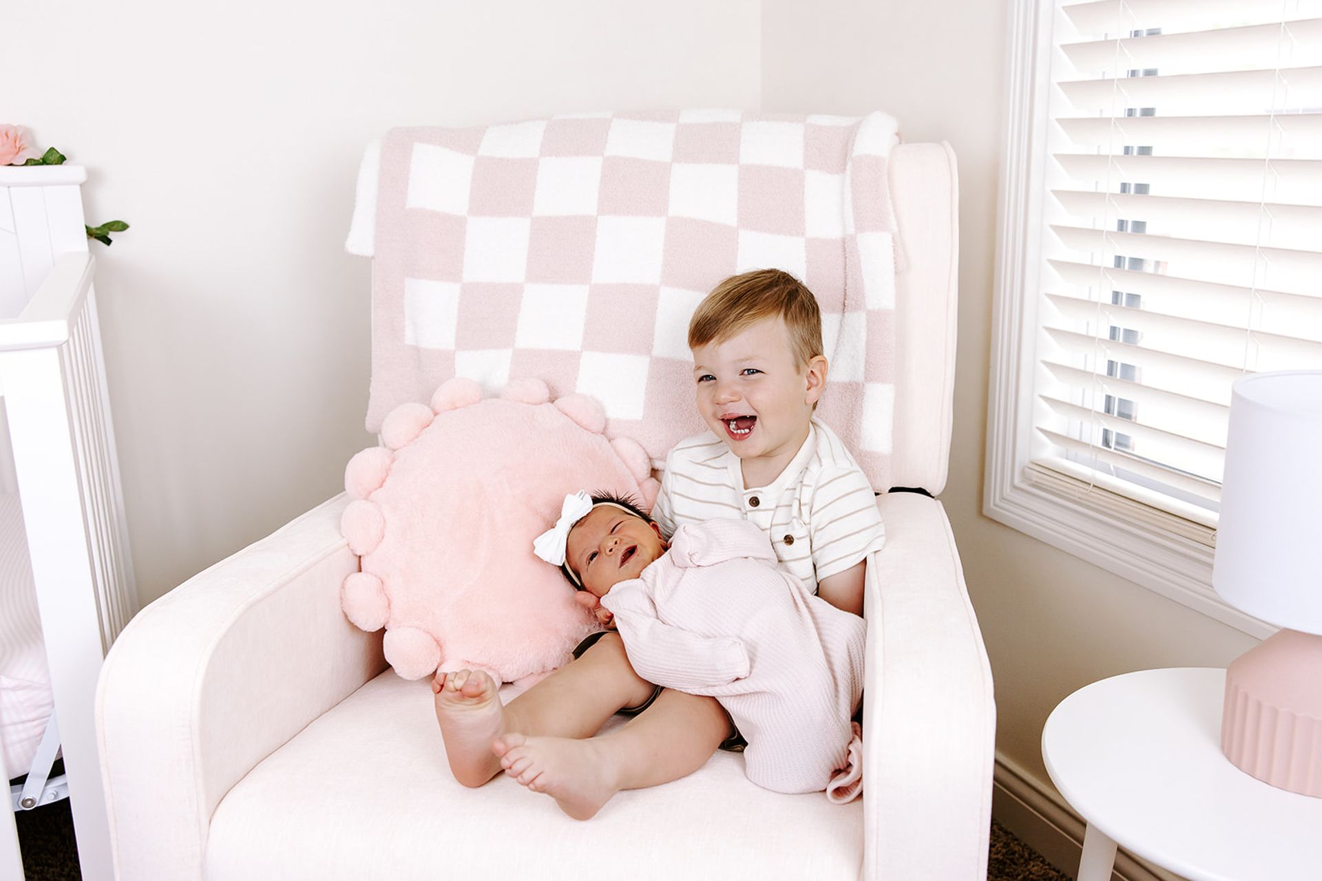 Boy laughing, holding baby in a pink nursery. Checkerboard blanket, pink accent pillows, white chair, and dresser.