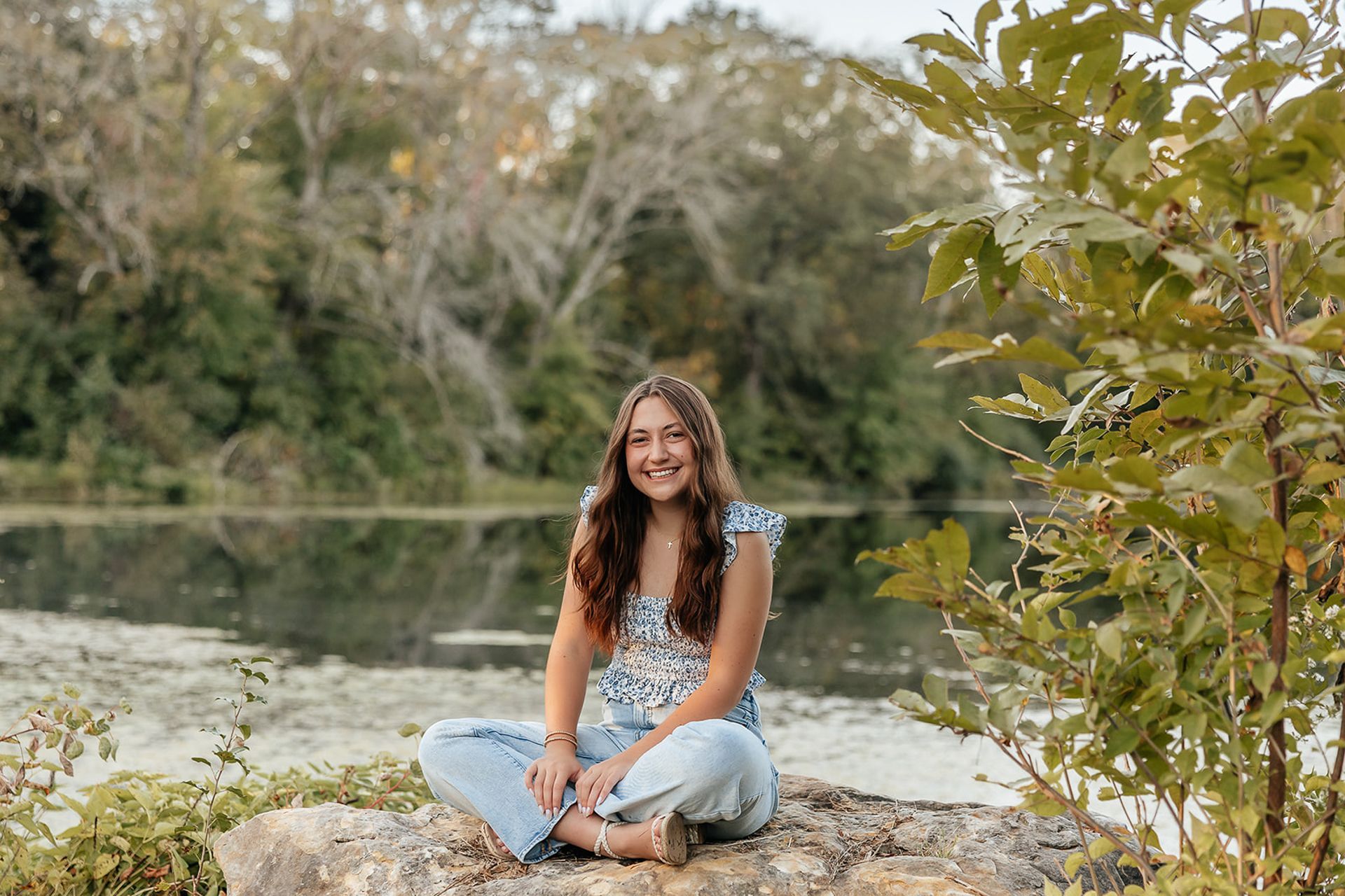 Woman sits on a rock by a lake, smiling. Wearing blue floral top and jeans. Outdoors with trees and water.