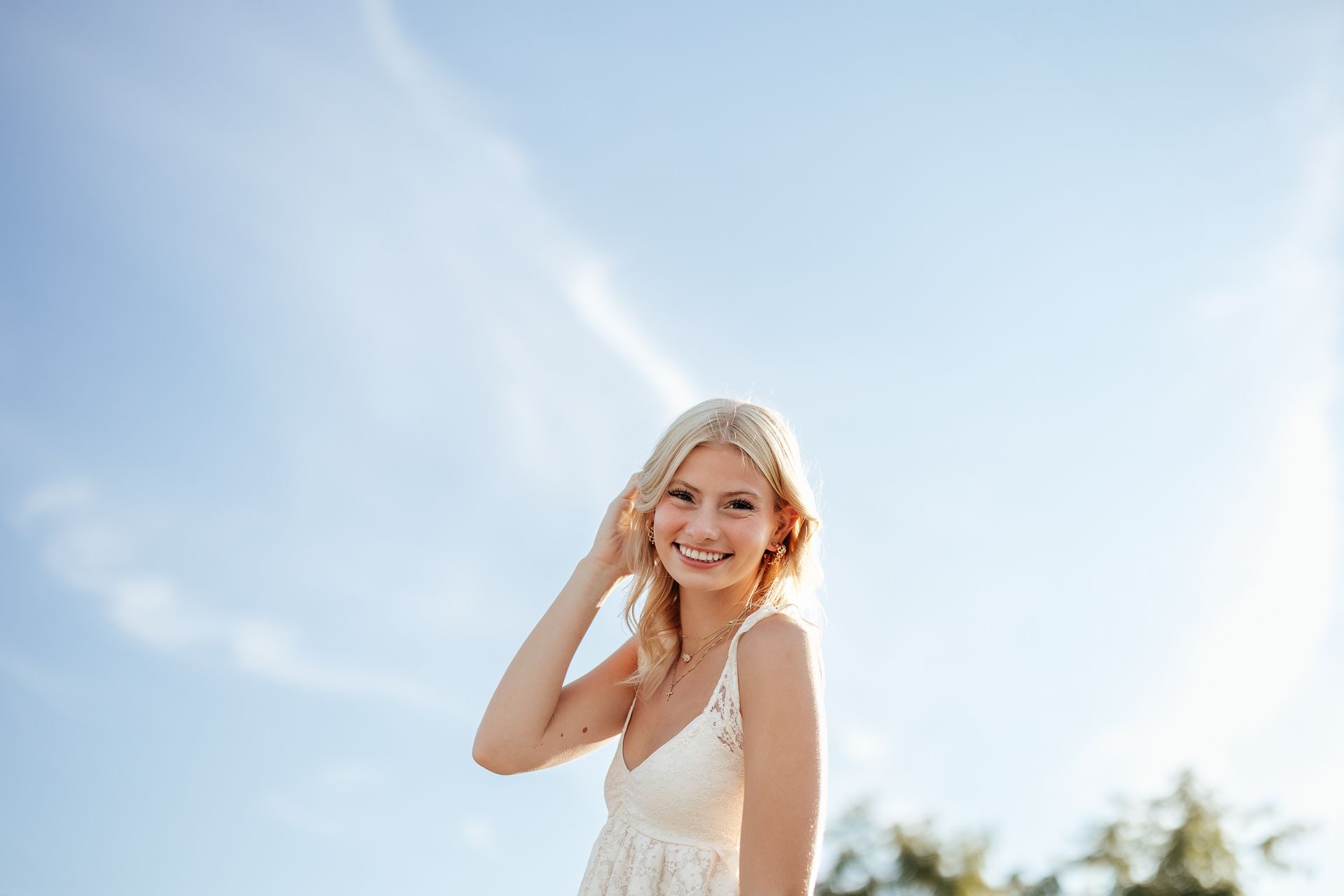 Woman smiling, touching her hair; blue sky background, wearing a white dress.