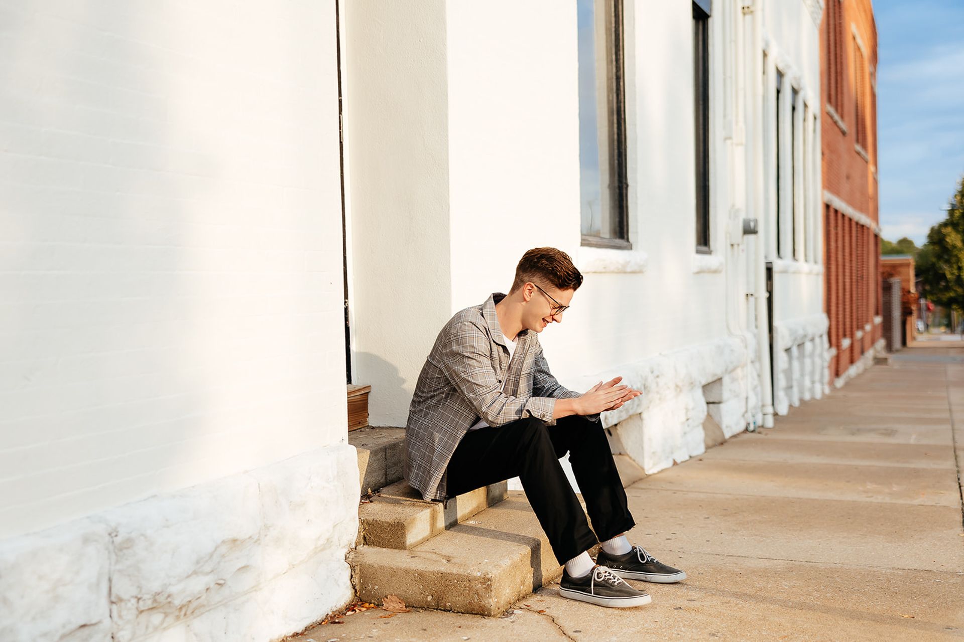 Person seated on steps of a white building, looking down, smiling. Wearing jacket, black pants, sneakers, outdoors.