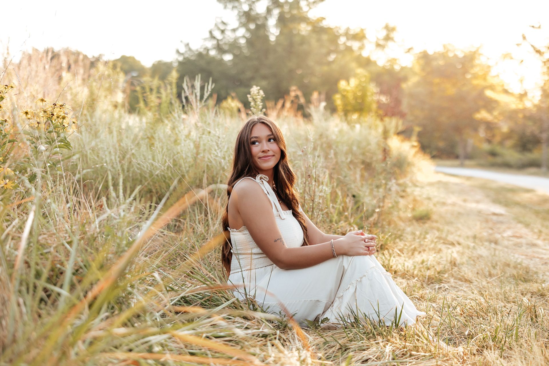 Woman in white dress sits in tall grass, looking to the side in a field bathed in sunlight.
