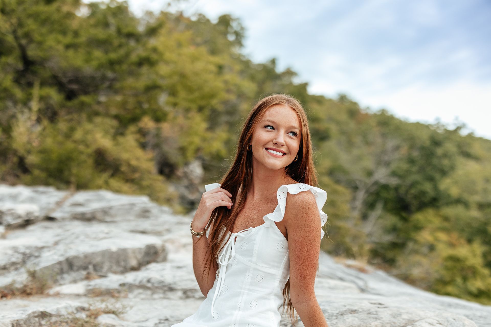 Woman with long red hair smiles, wearing white dress, on rocky landscape with trees.