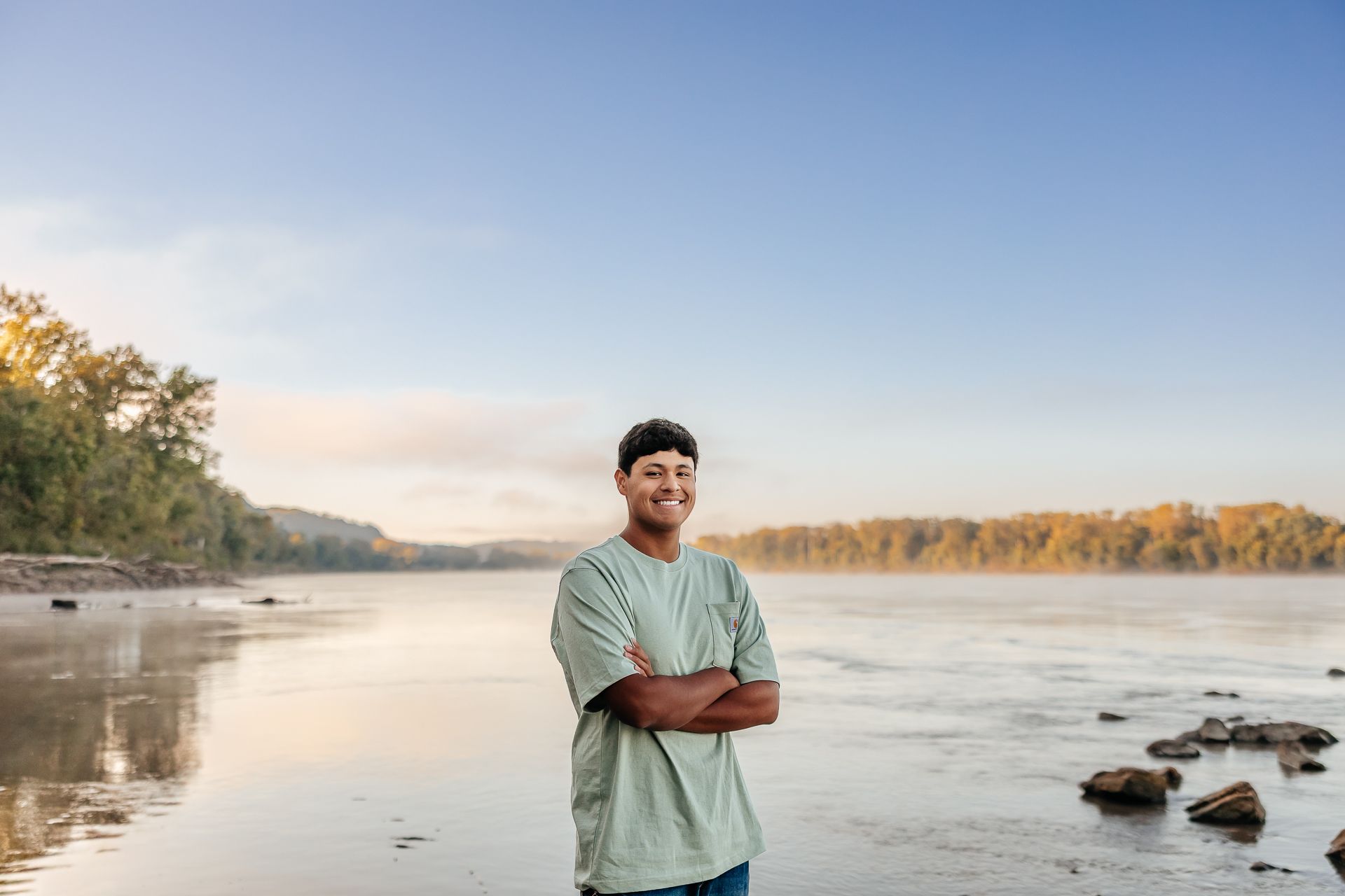 Man stands arms crossed, smiling, near a misty river with a shoreline of rocks and trees. Blue sky.