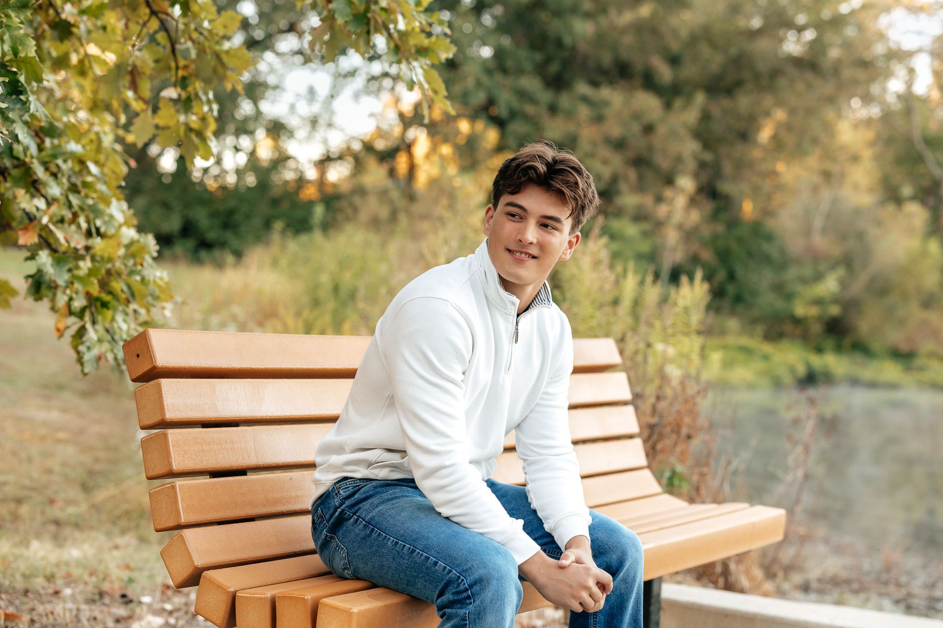 Man sitting on a park bench, looking to the side with a smile. Outdoors, wearing a white sweater and jeans.