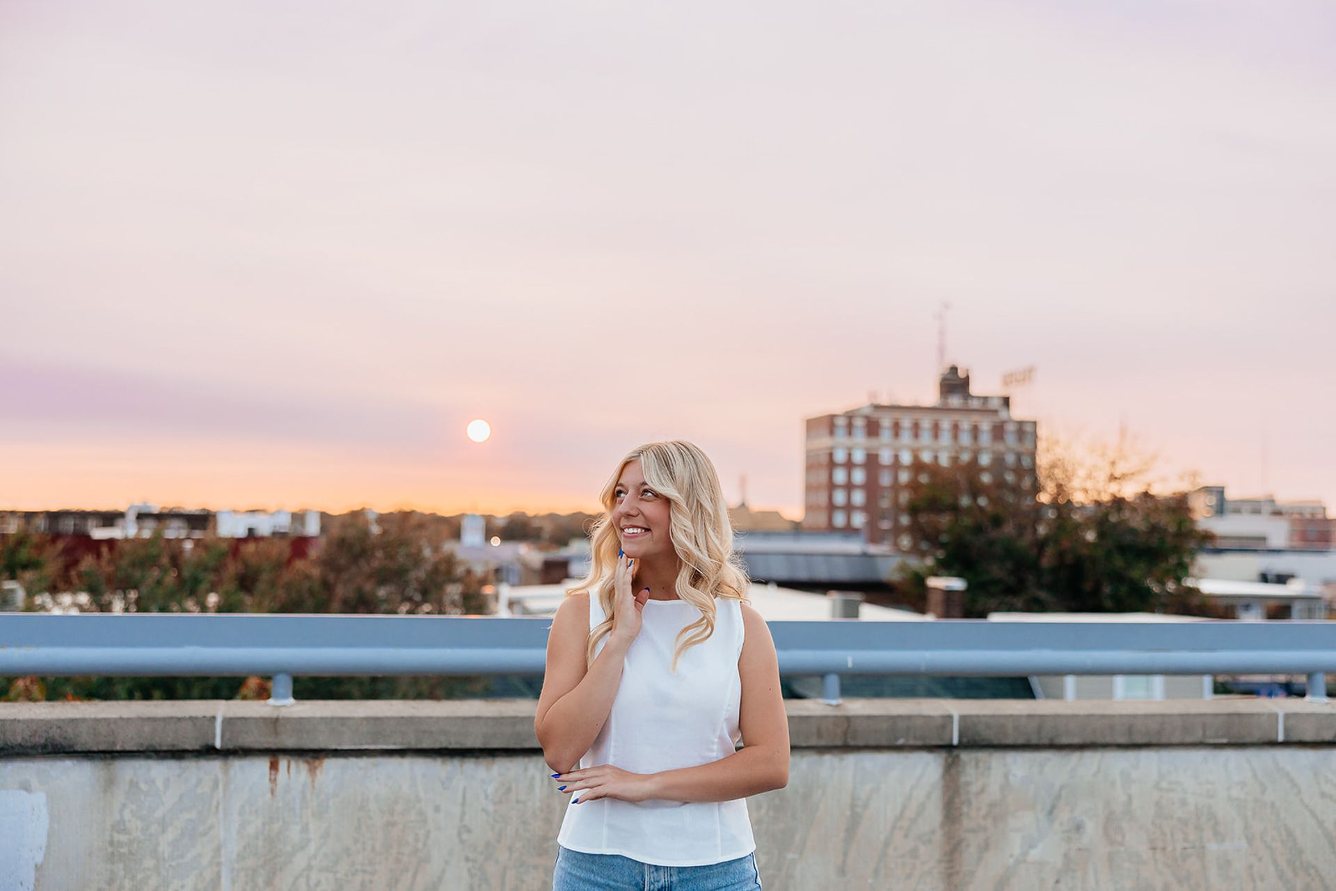 Blonde woman smiles, resting chin on hand, on a rooftop at sunset, city in background.